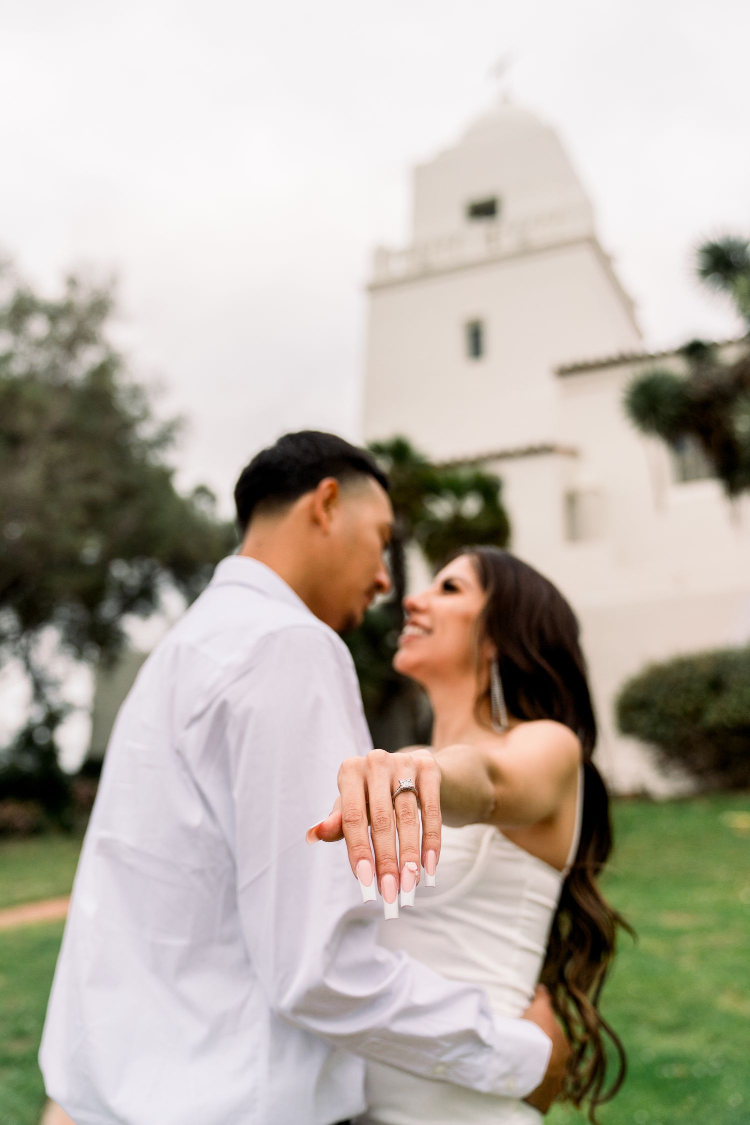 A newly engaged couple shows off ring while embracing in front of a white Spanish mission-style church tower.