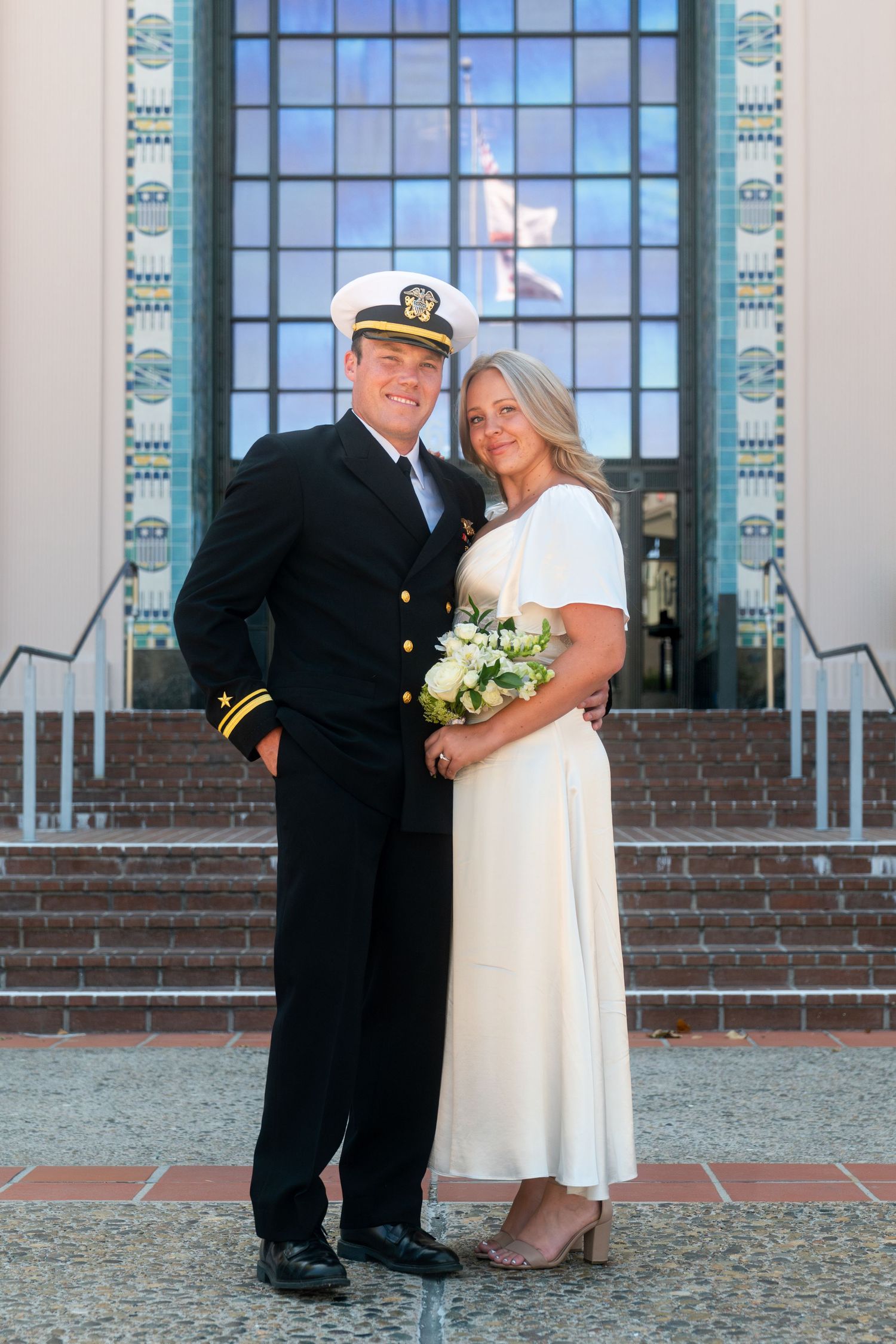 A couple in wedding attire sharing a romantic moment in front of a brick building with large arched windows.