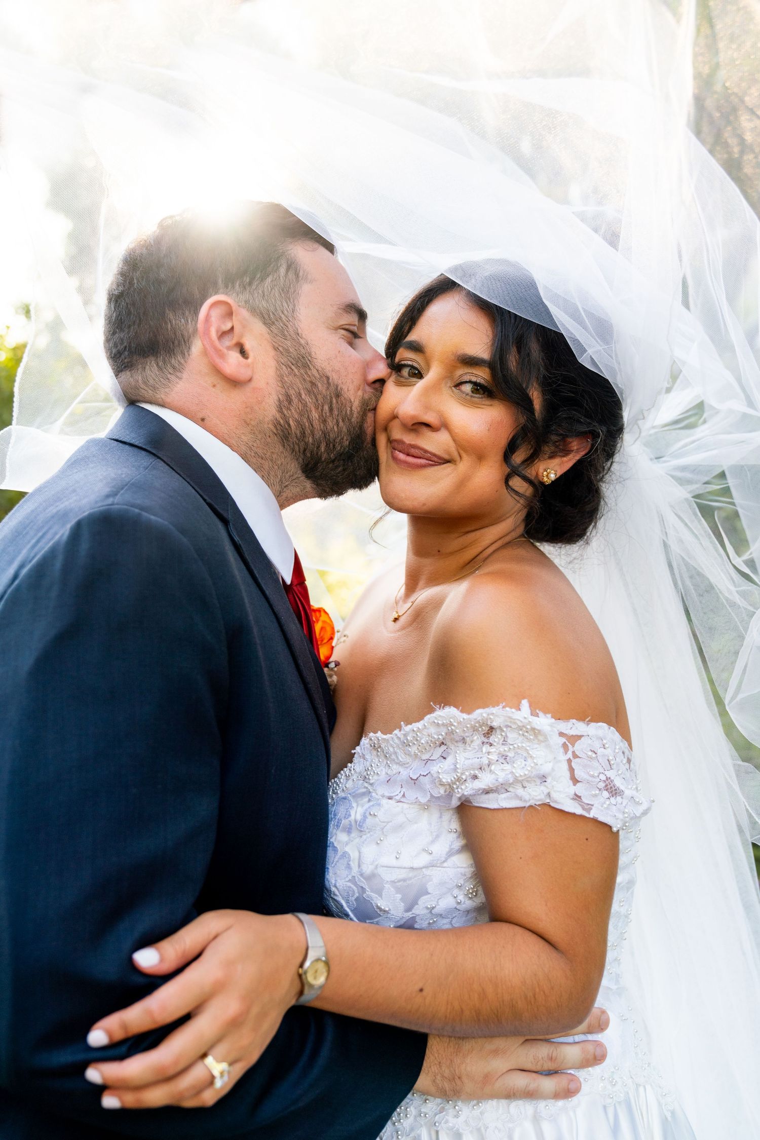 A tender moment between newlyweds as they embrace during their outdoor wedding, with sunlight and flowing white veil in background.