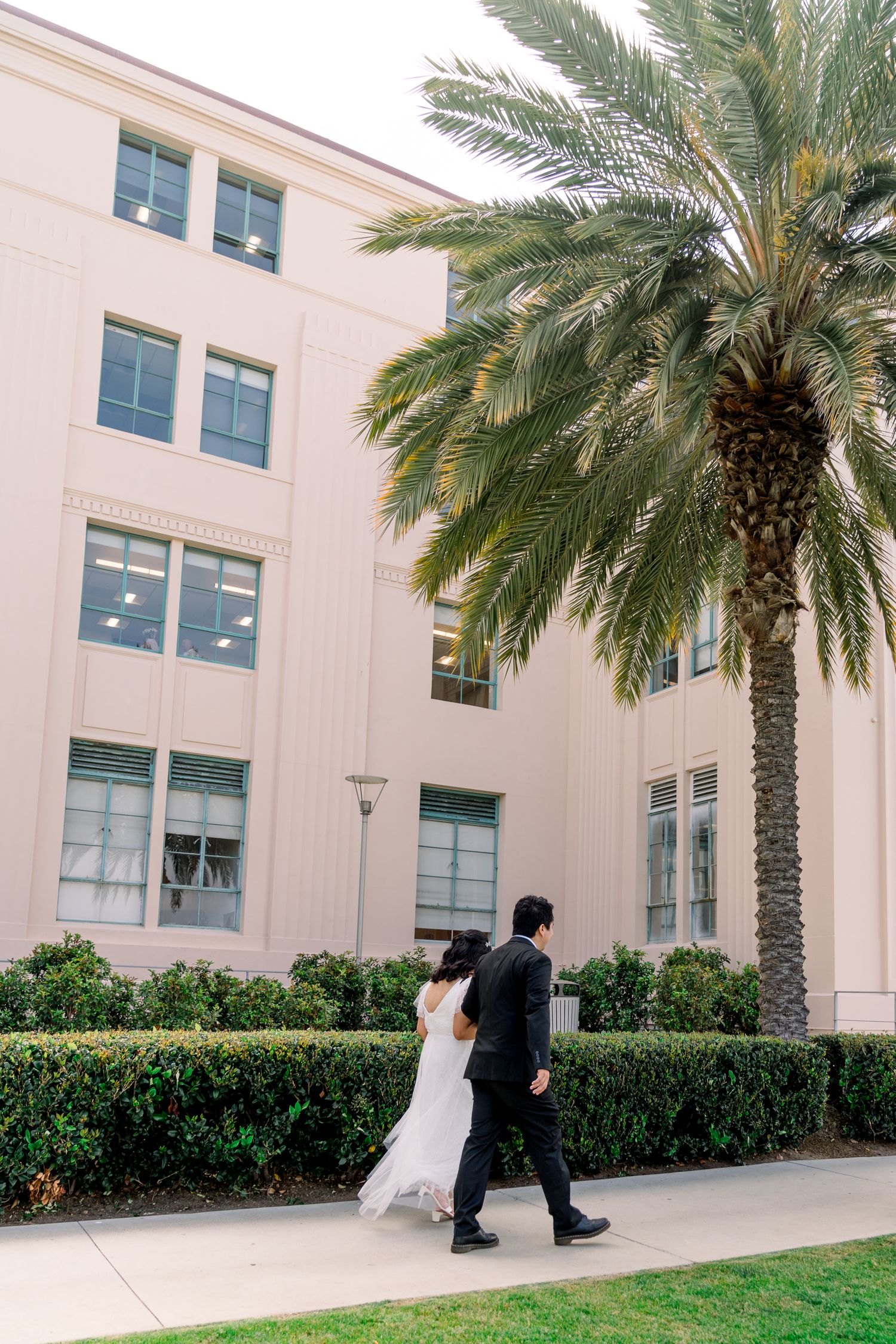 A couple walks along a sidewalk next to a pink building with a tall palm tree on a sunny day.