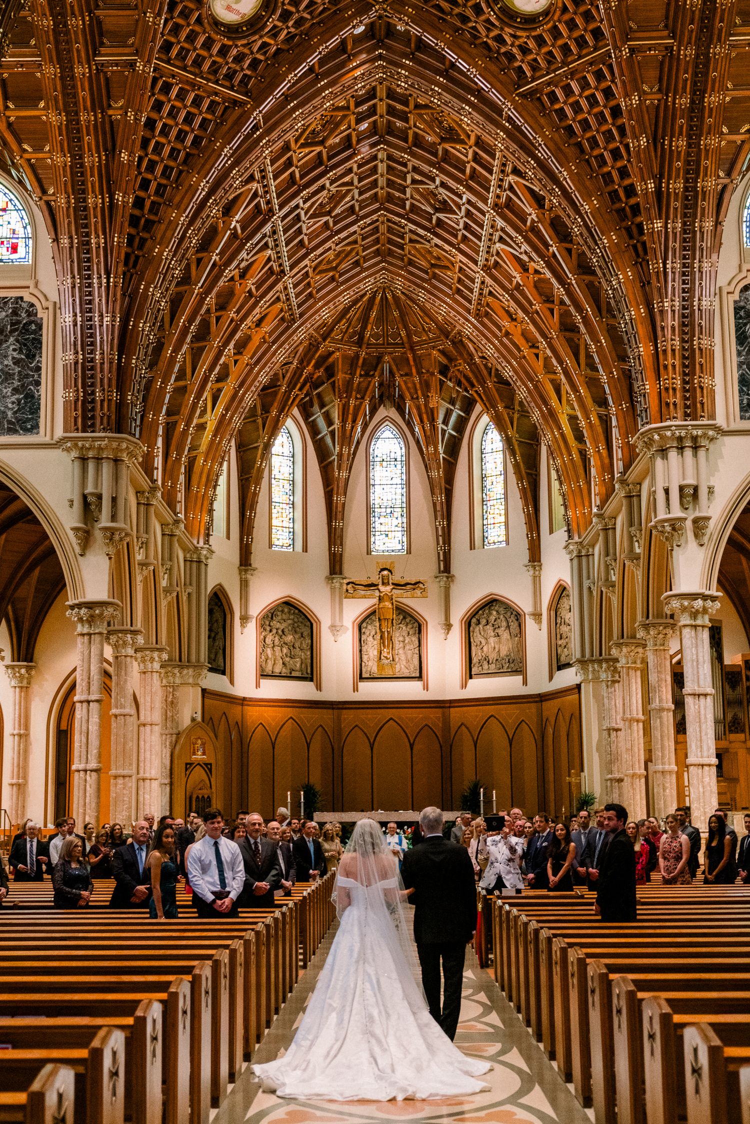 A cathedral wedding ceremony takes place under soaring Gothic arches with ornate wooden vaulted ceilings and stained glass windows.