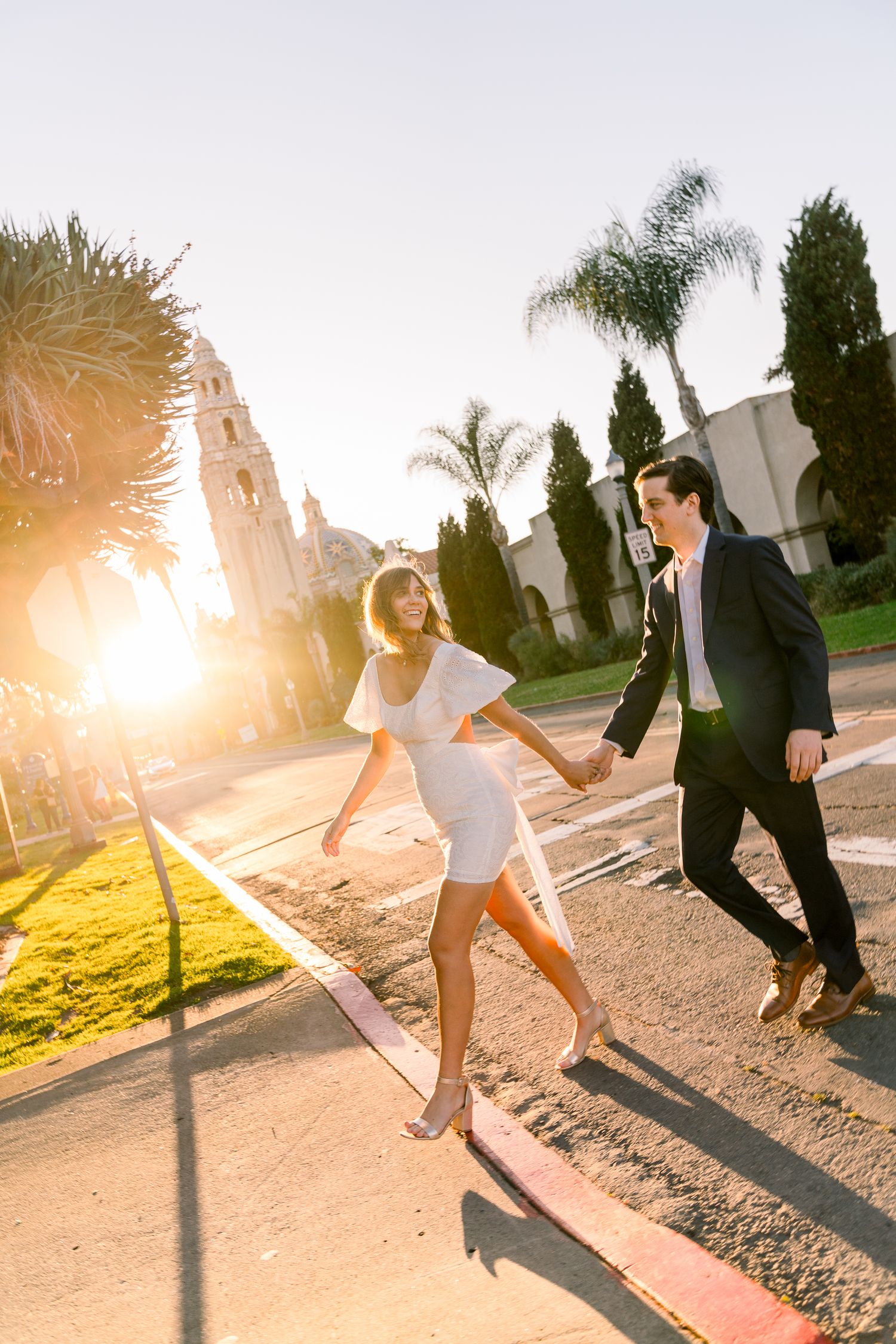 Couple holding hands and walking along a palm-lined street during golden hour sunset with dramatic lens flare.