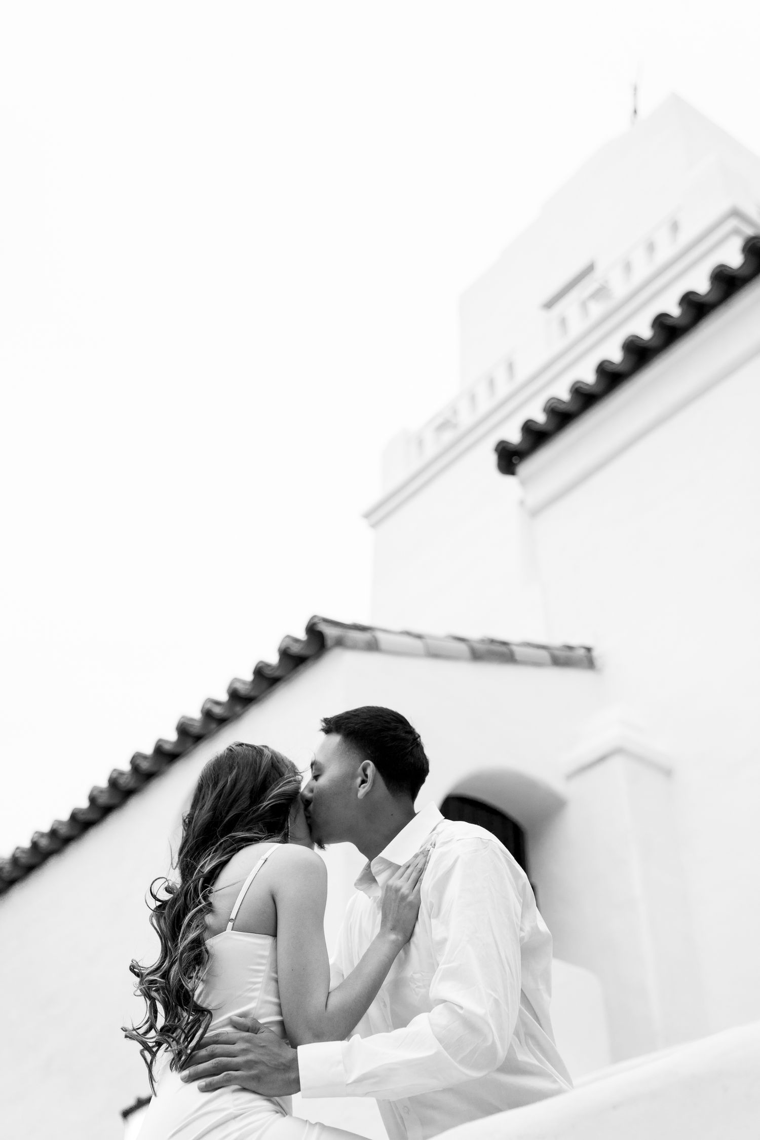A romantic black and white photo of a couple kissing against a white Mediterranean-style building with tiled roof.