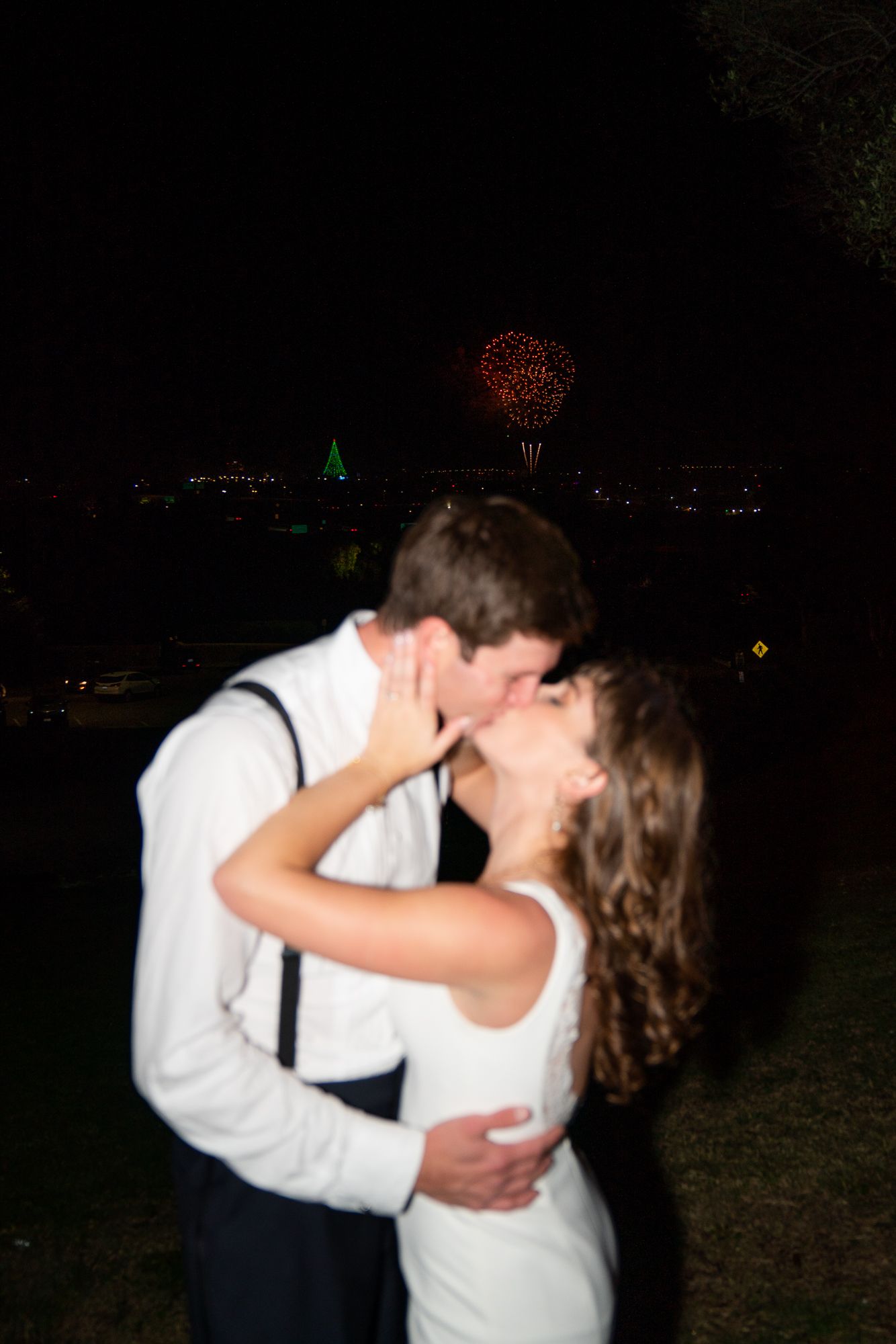 A romantic couple kissing at night with fireworks bursting in the dark sky over a city skyline.