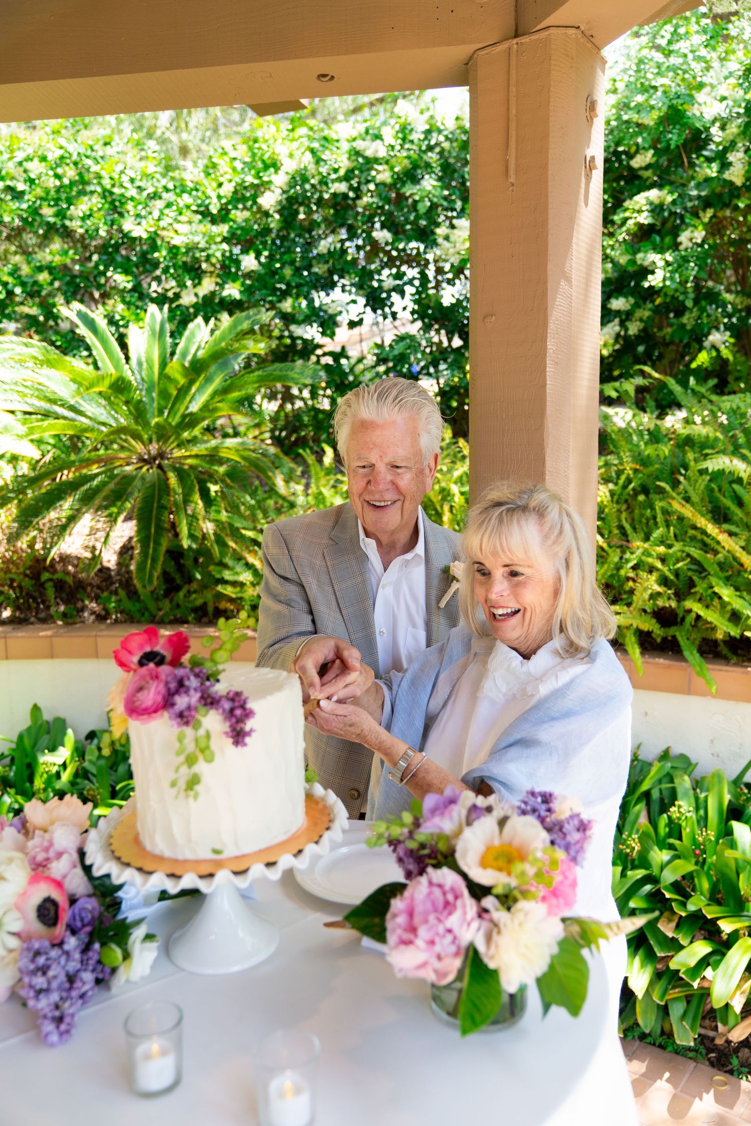 A couple shares a joyful moment cutting into an elegant white wedding cake adorned with fresh flowers on a patio.