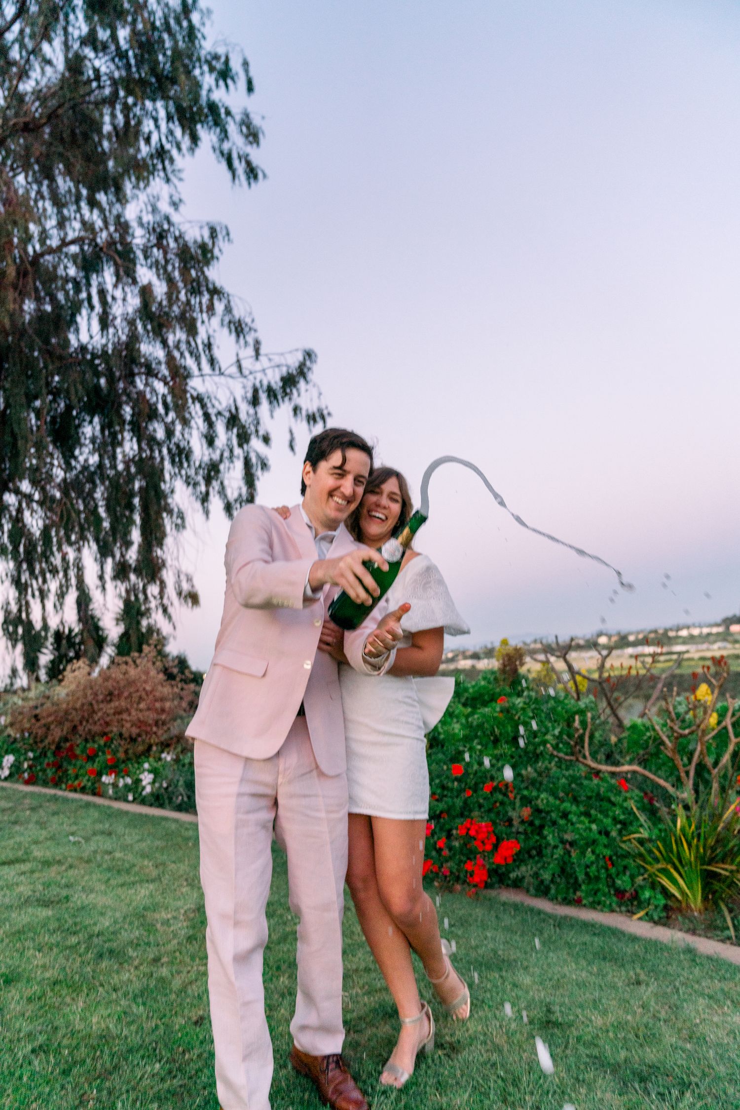 A couple pops champagne while celebrating on a scenic lawn with flowers and trees at sunset.