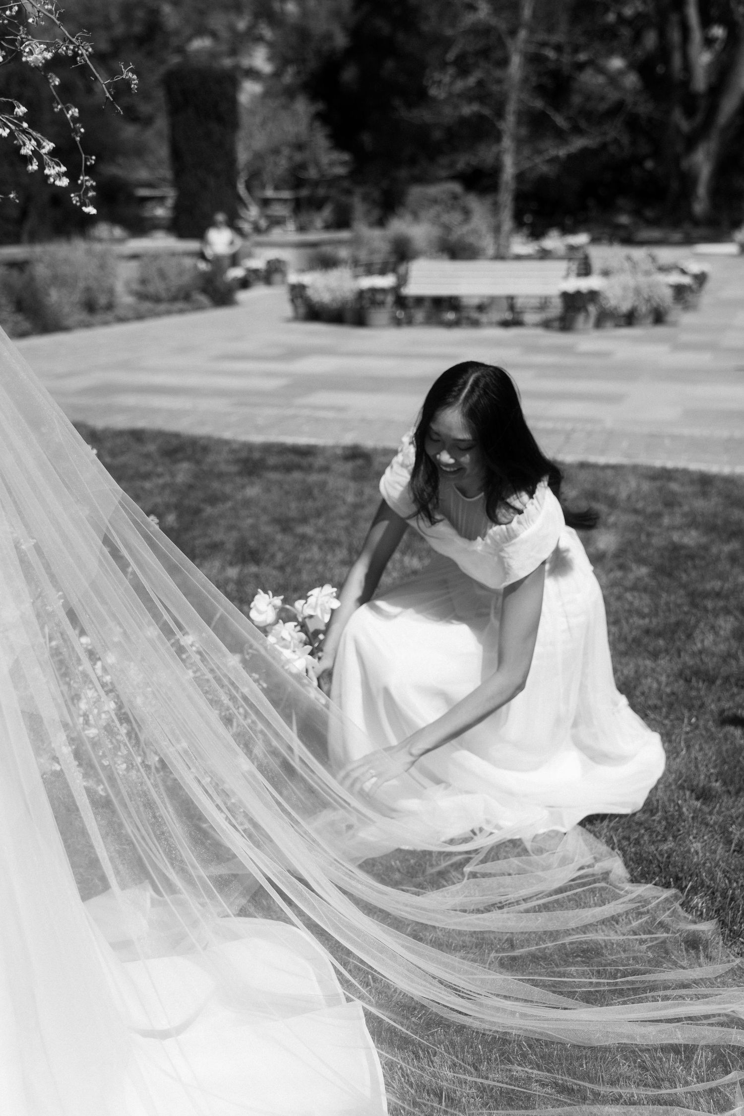 A bride in a white dress sits on grass while adjusting her long flowing veil in a garden setting.