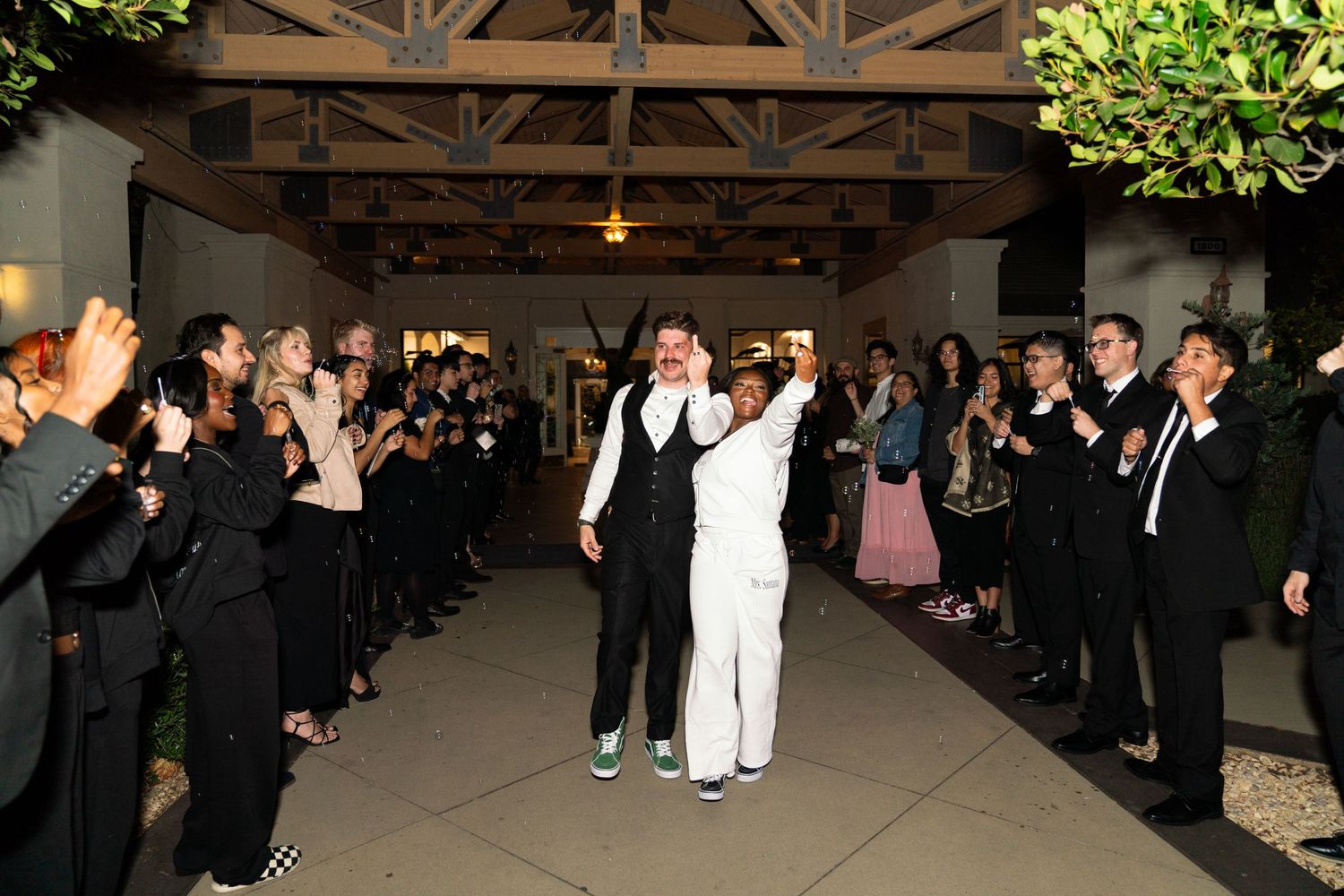 Newlyweds celebrate their first dance together while guests line the sides of an indoor venue cheering and taking photos.
