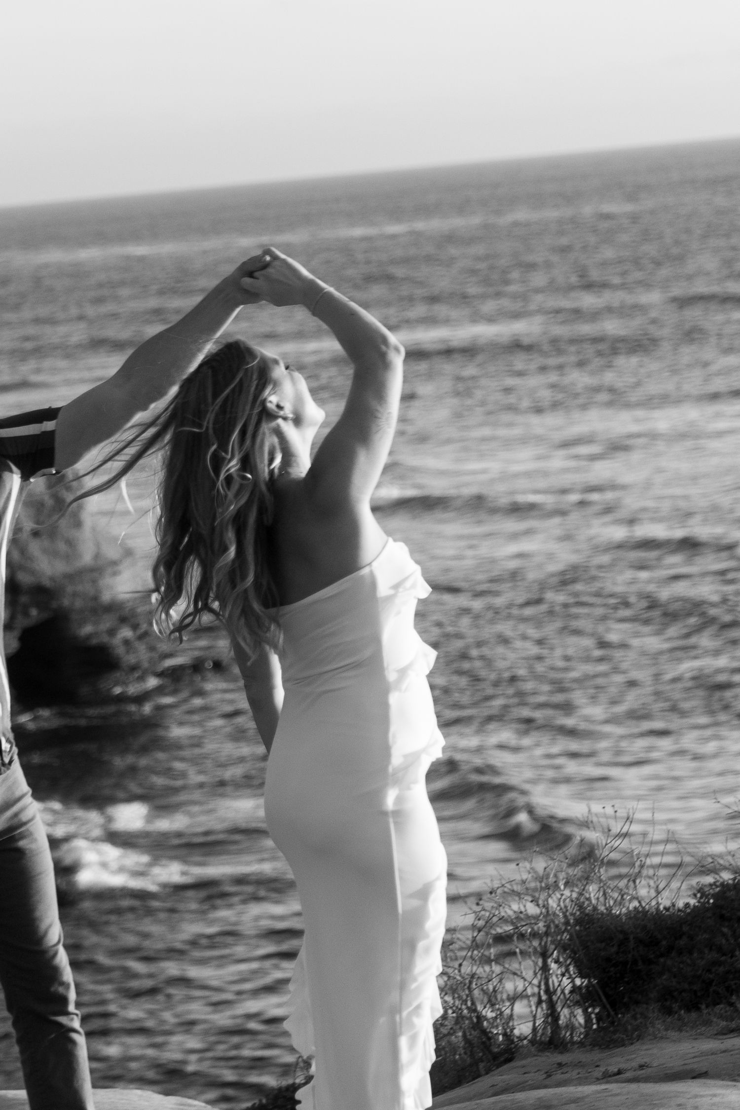 A person in a white dress dances gracefully against an ocean backdrop in black and white photography.