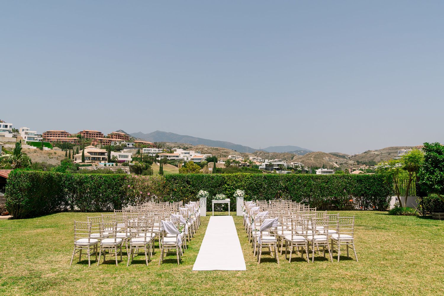 Outdoor wedding ceremony setup with white chairs and aisle runner on green lawn overlooking hillside Mediterranean village.