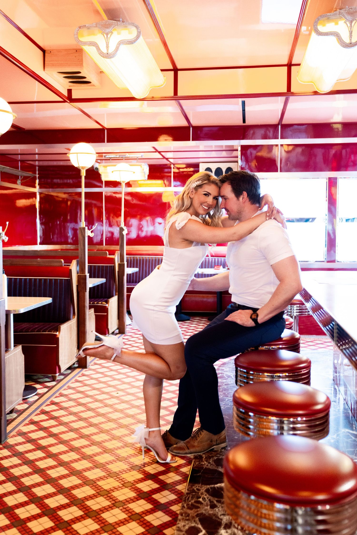 A romantic retro diner scene with red booths, vintage barstools, and checkered flooring under warm lighting.