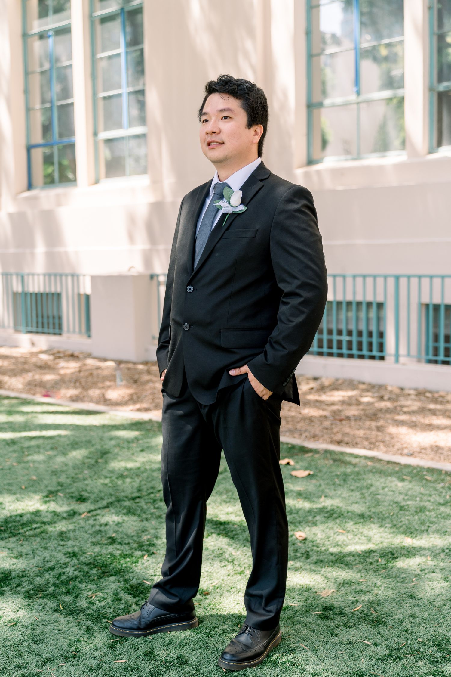 Professional photo of a person in a dark suit and blue tie standing on grass in front of a building with hands in pockets.