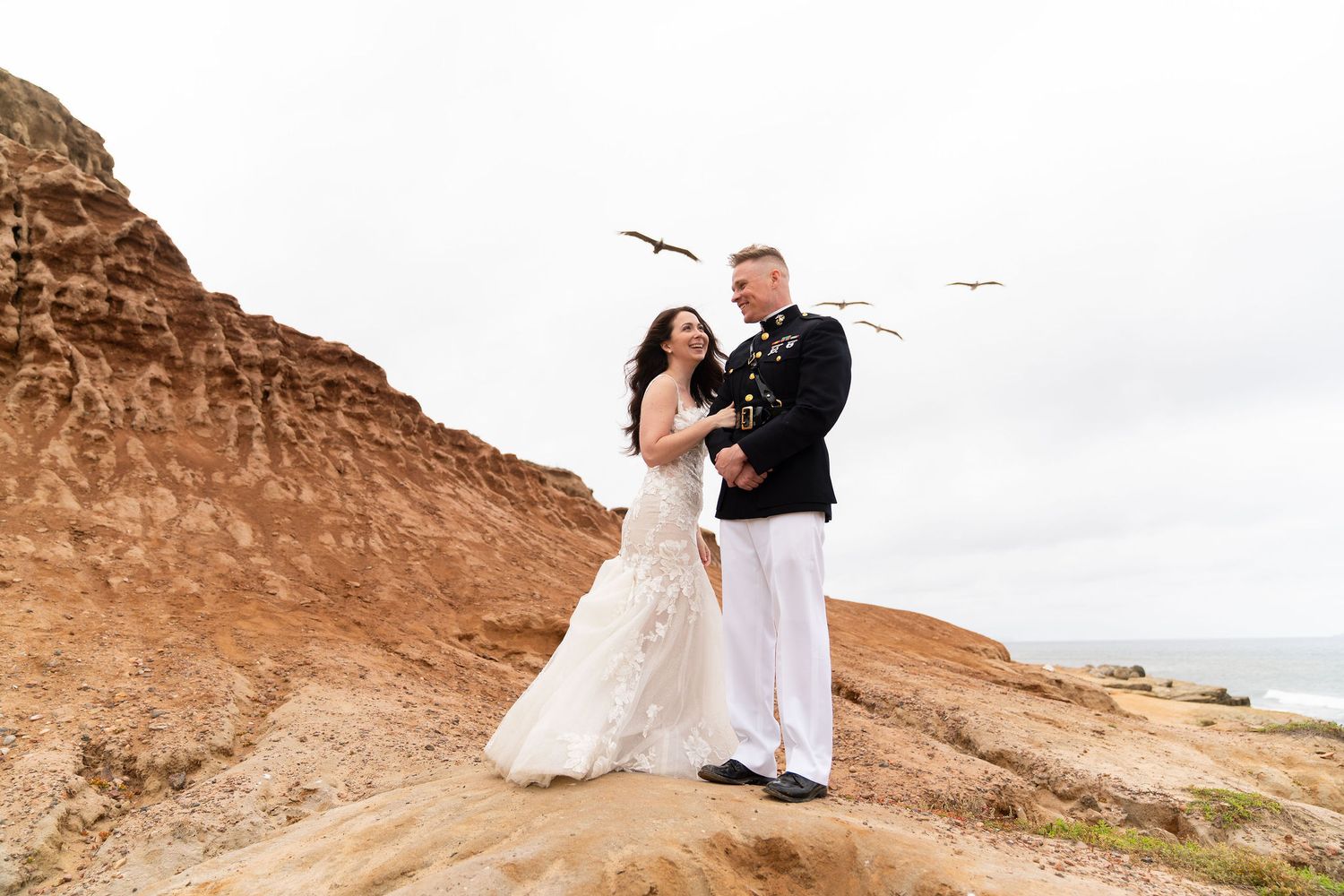 Two people embrace against a backdrop of rocky desert cliffs and mountains during a sunny outdoor photo session.