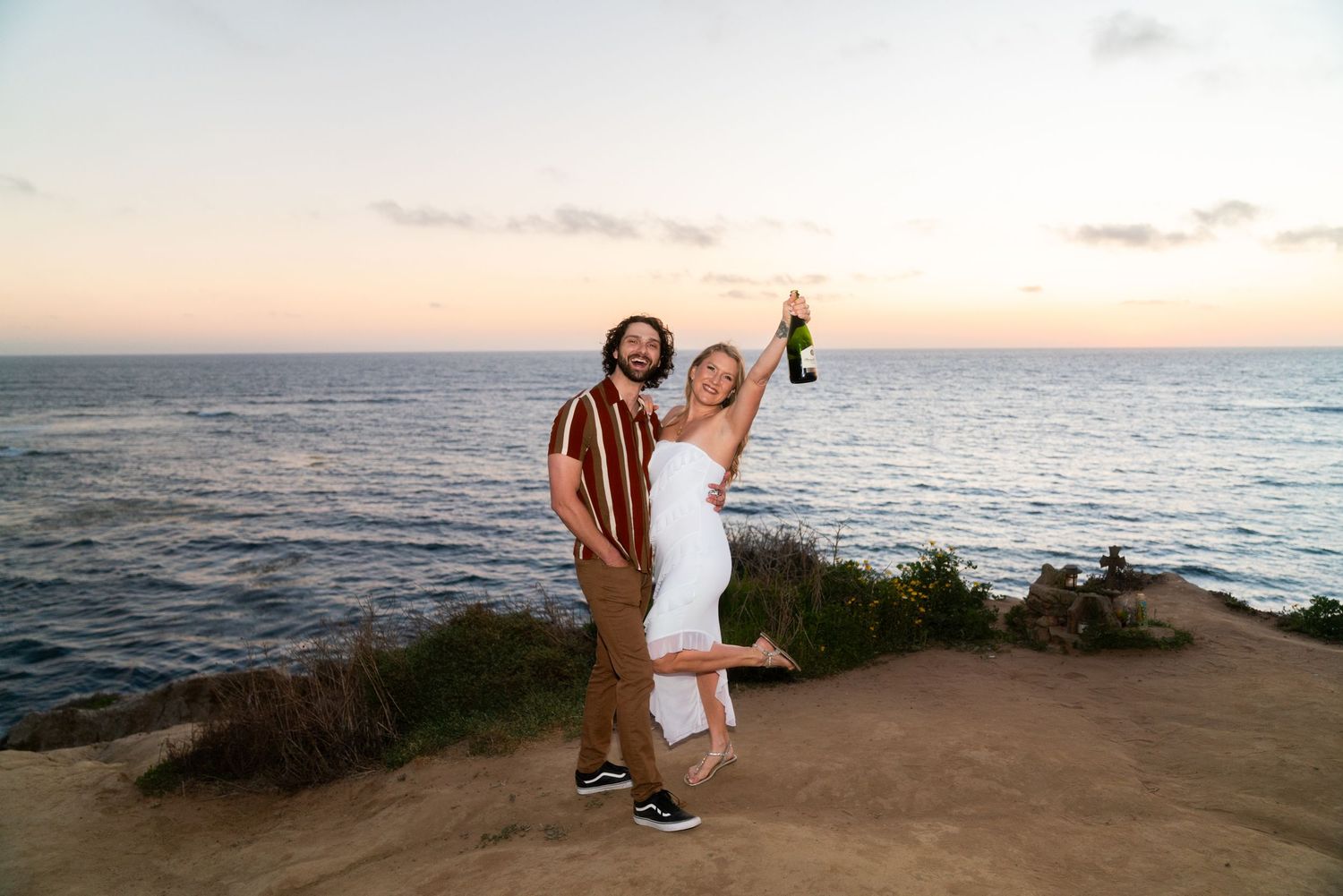 Two people celebrate joyfully on a coastal cliff overlooking the ocean at sunset.