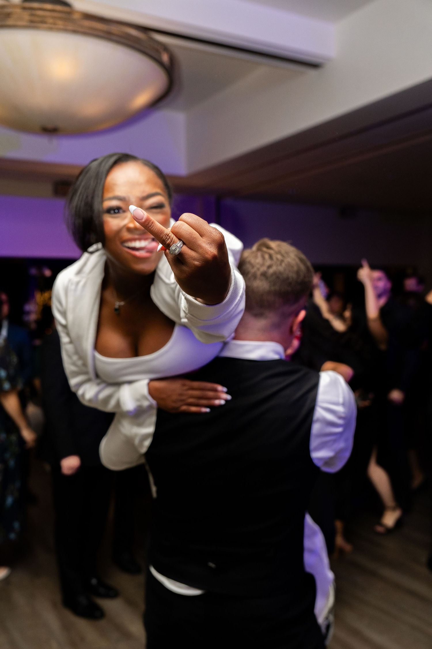 At a party reception, someone in a white blazer leans forward with a joyful expression while dancing with others.