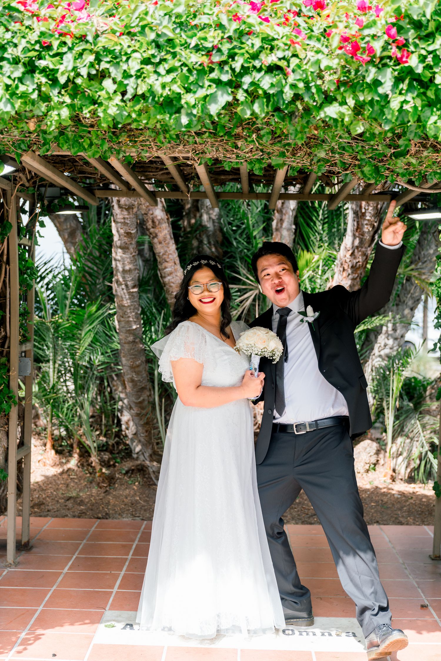 A joyful couple celebrates under a rustic wooden pergola draped with pink flowers and greenery during their garden wedding.