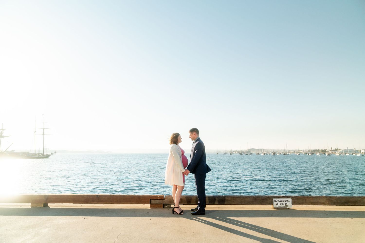 A couple stands together on a pier overlooking a tranquil waterfront on a bright sunny day.