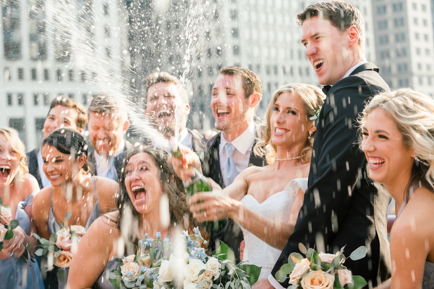 Wedding party members laugh joyfully as they are sprayed with champagne during outdoor celebration.