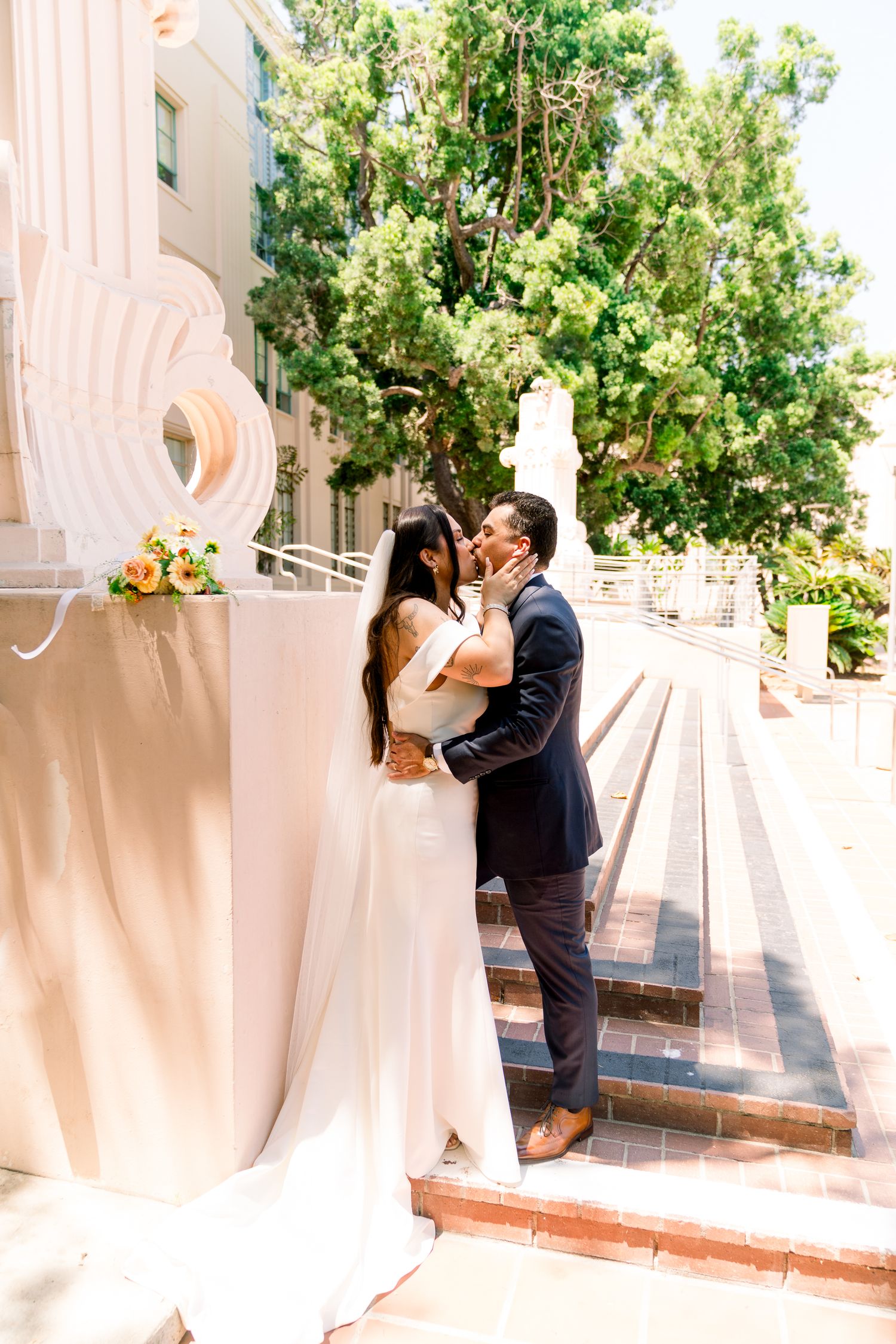 A couple shares a romantic kiss on outdoor steps of an elegant venue with greenery and sunshine in the background.