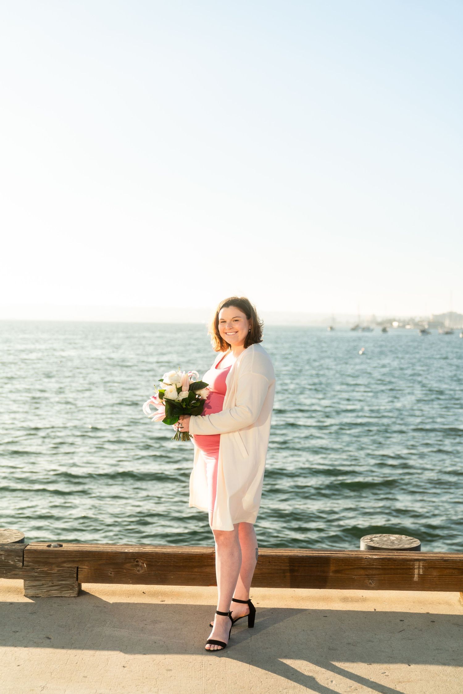 A person in a coral dress and white cardigan holds flowers while standing on a pier by the ocean at sunset.