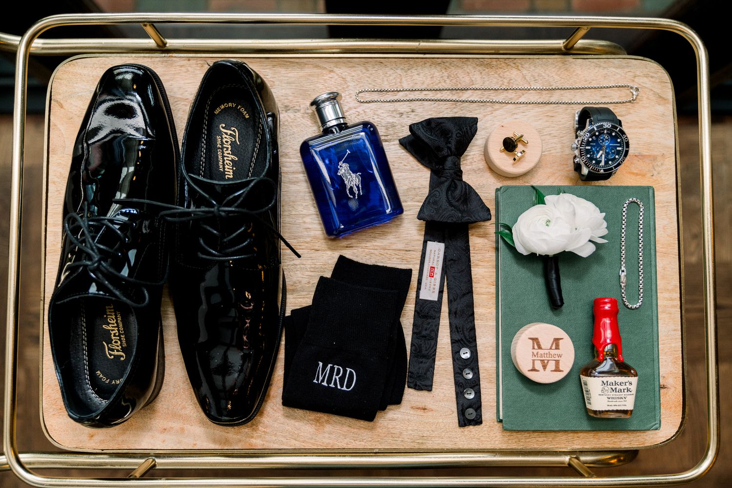 Overhead view of men's grooming accessories including black dress shoes, cologne, watch, and bow tie on wooden surface.