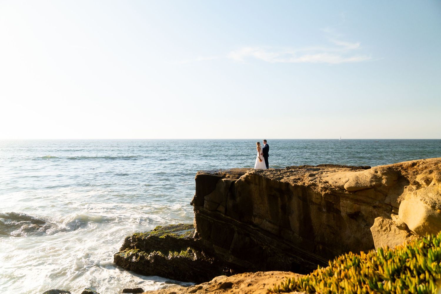 A couple stands on a coastal cliff overlooking the Pacific Ocean with waves crashing against rocky shores in California.