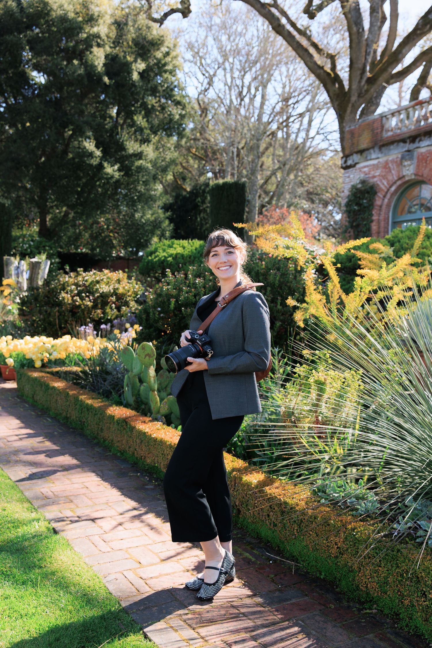 A photographer standing on a brick path in a garden with yellow tulips and ornamental grasses on a sunny day.