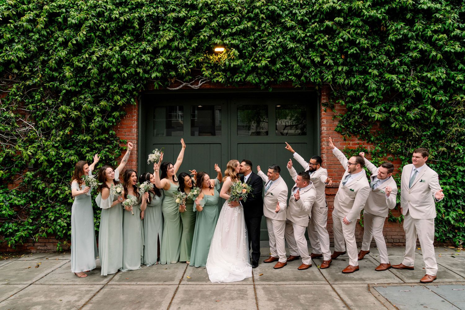 Wedding party throws their hands up in celebration in front of ivy covered wall during outdoor summer celebration.