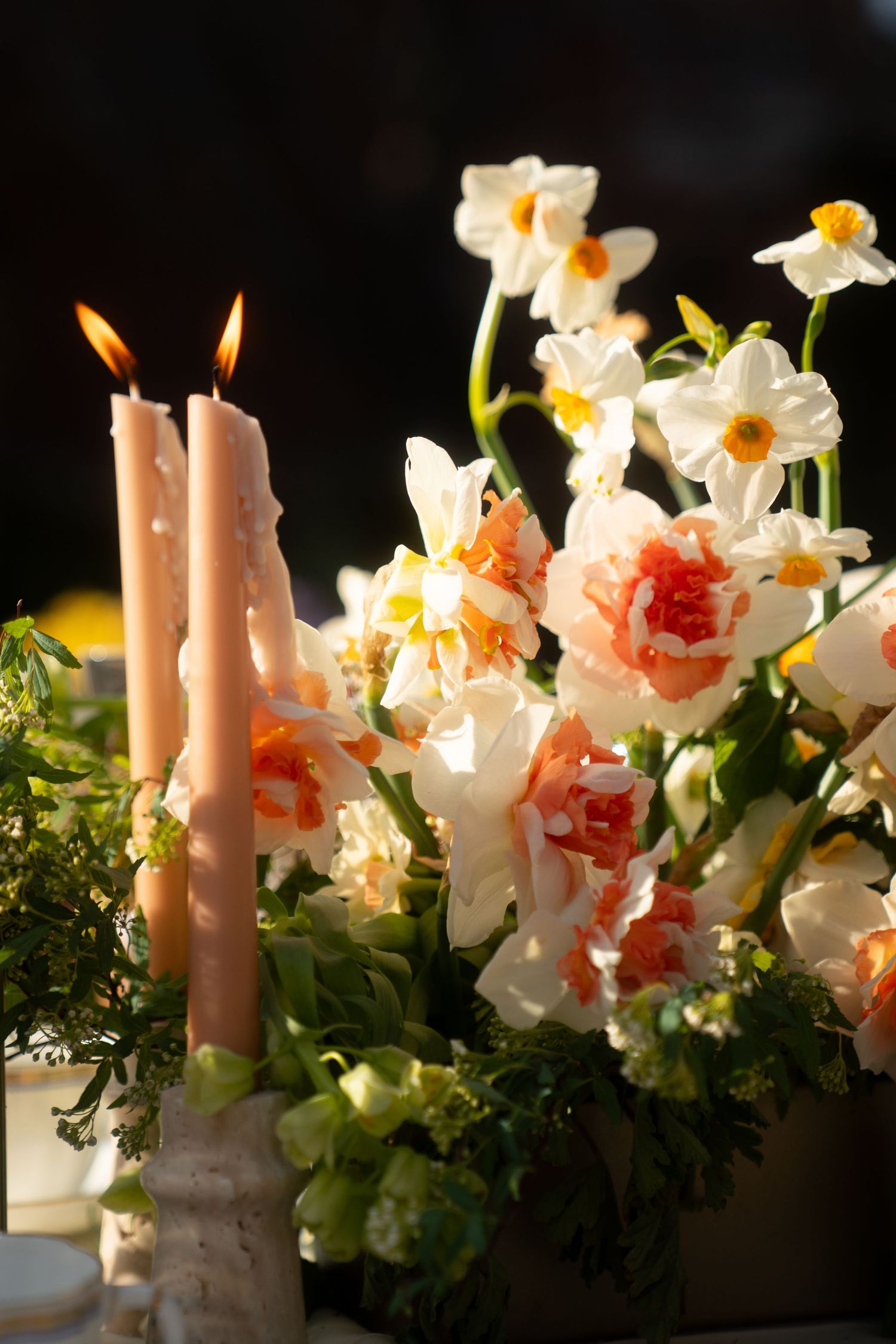 Pink lit candles glow beside delicate white and peach daffodils and roses in an elegant floral arrangement.