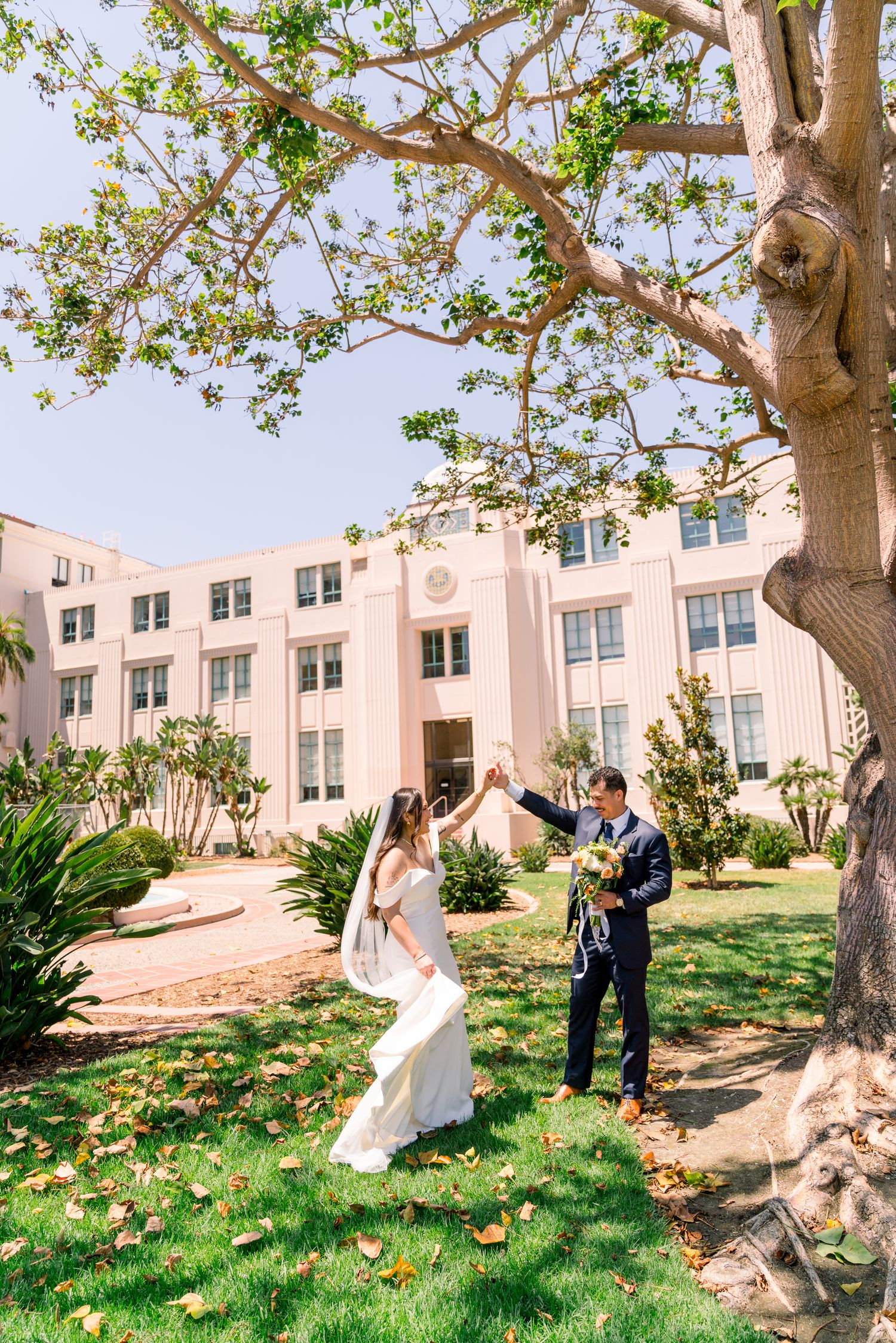 Couple celebrating their wedding day under a large tree outside a pink Art Deco style building on a sunny afternoon.