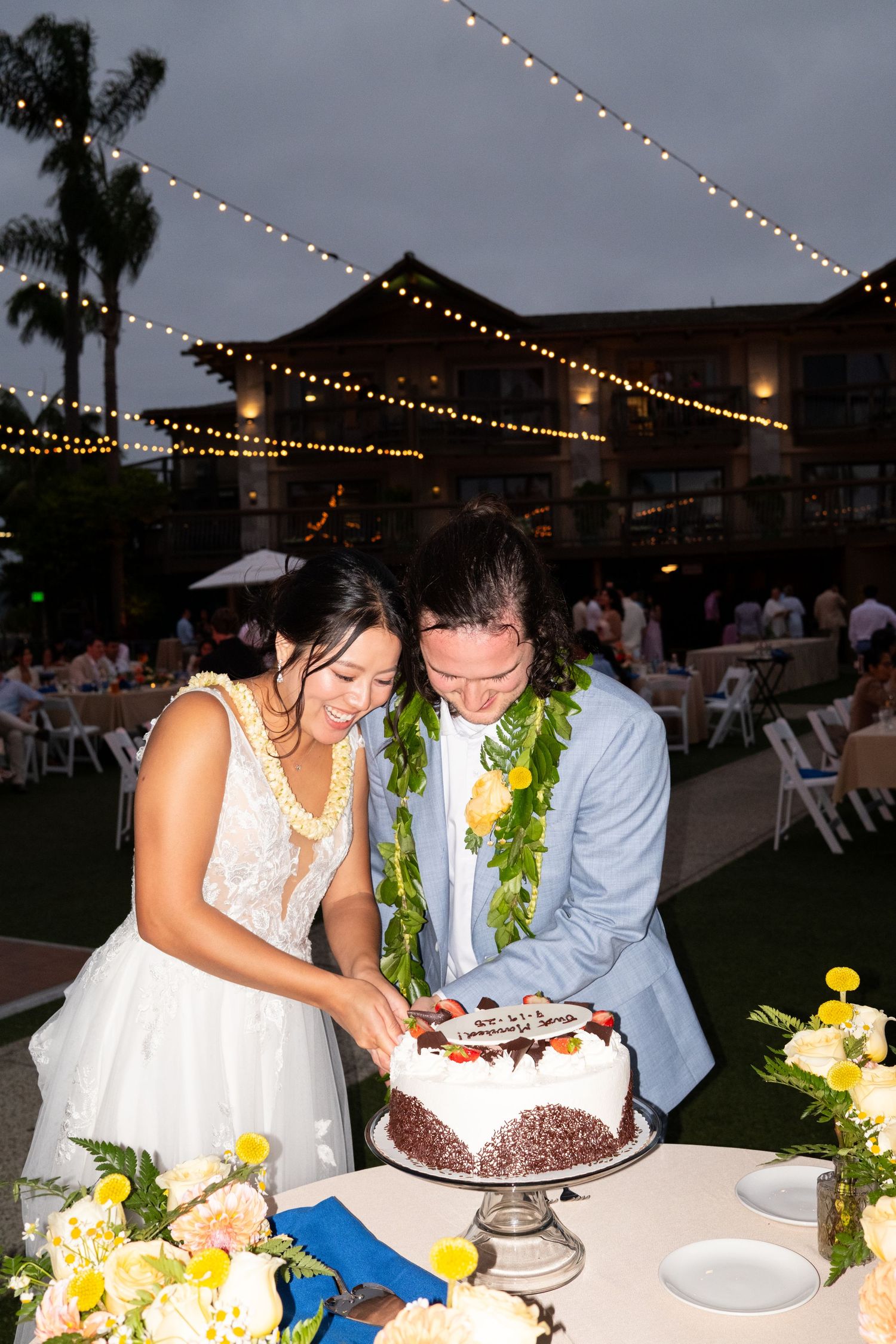 A couple in formal attire cuts their wedding cake together under twinkling string lights at an outdoor evening reception.