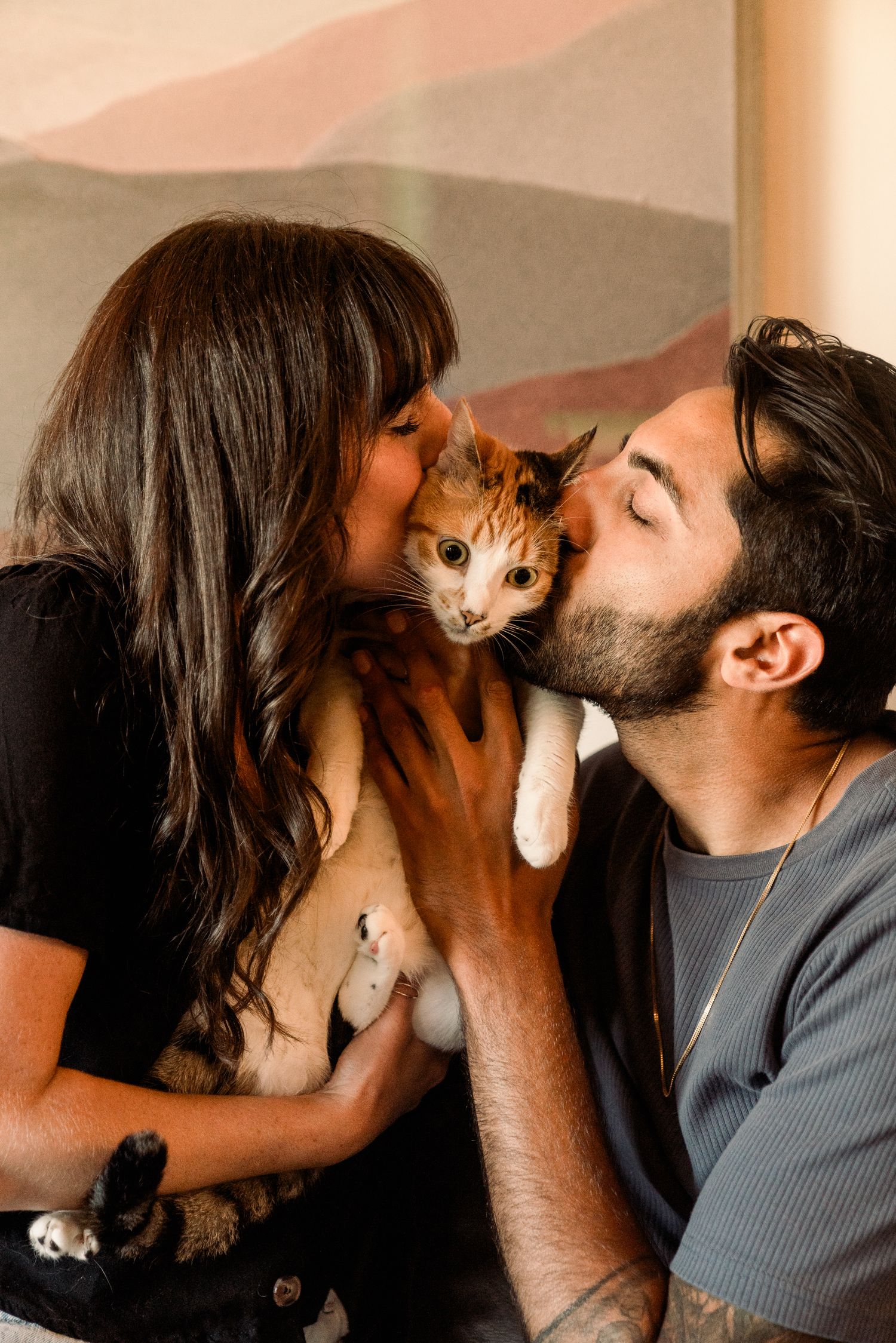 A couple sharing a tender moment while kissing their ginger and white cat held between them.