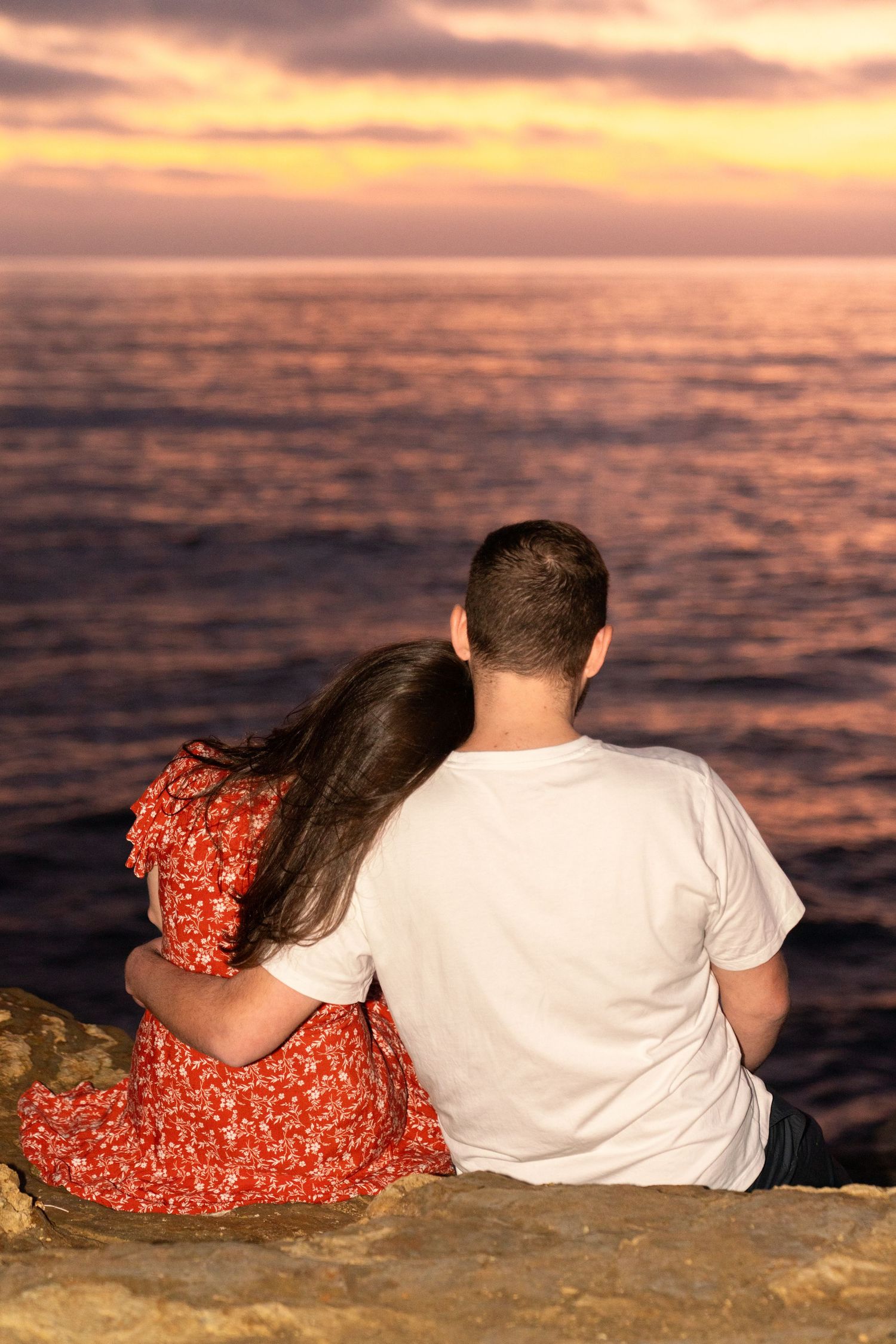 A couple sits together on a rocky shore watching the sunset over the ocean, silhouetted against an orange sky.
