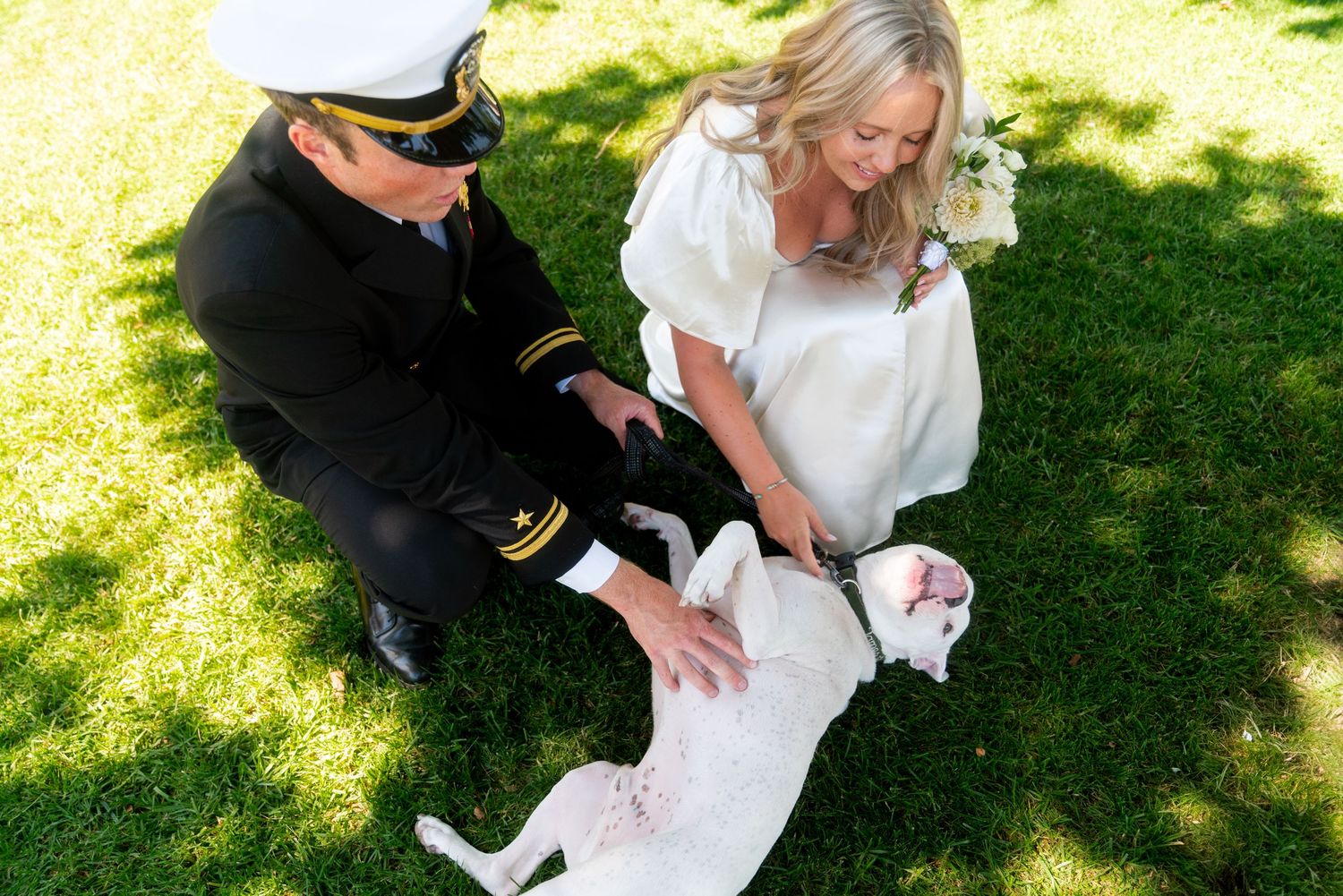 Two people in a sunny garden petting a happy white dog lying on the grass.