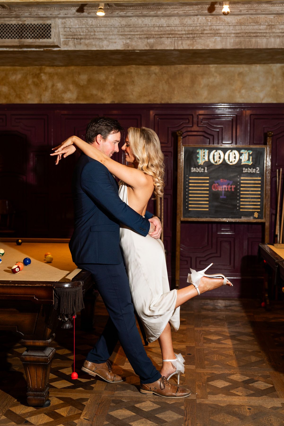 A couple dancing in formal attire next to a pool table with a vintage jukebox in the background.