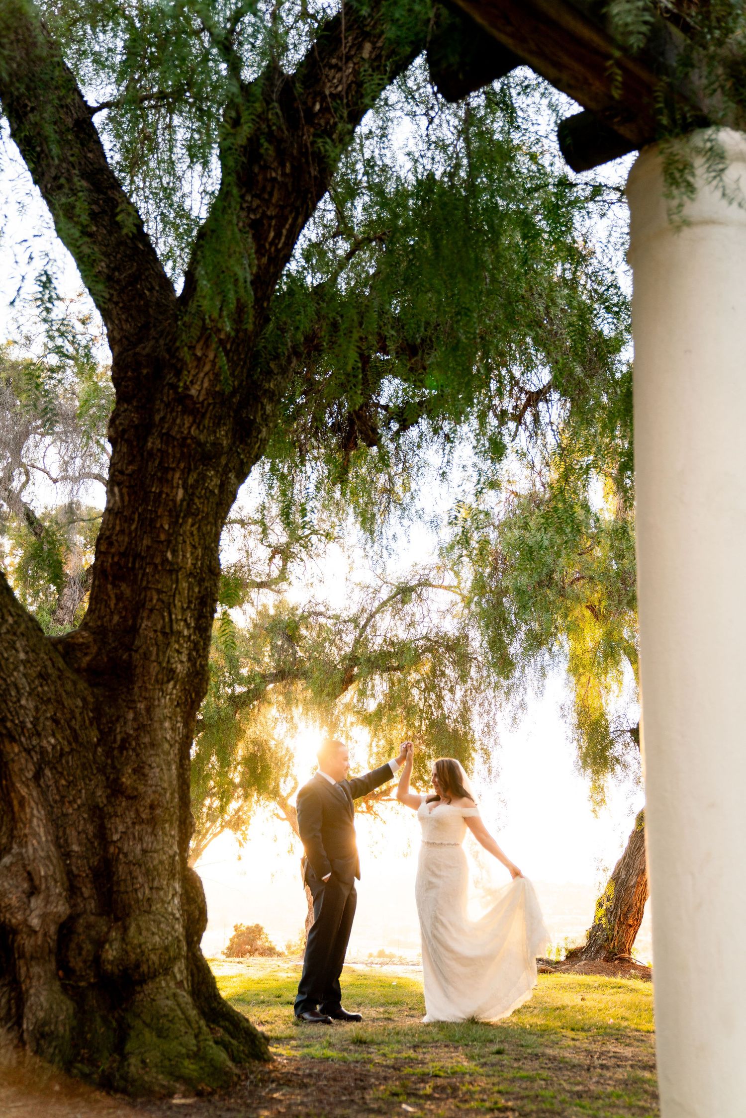 A couple shares a romantic moment under a large weeping willow tree with sunlight streaming through the branches at sunset.