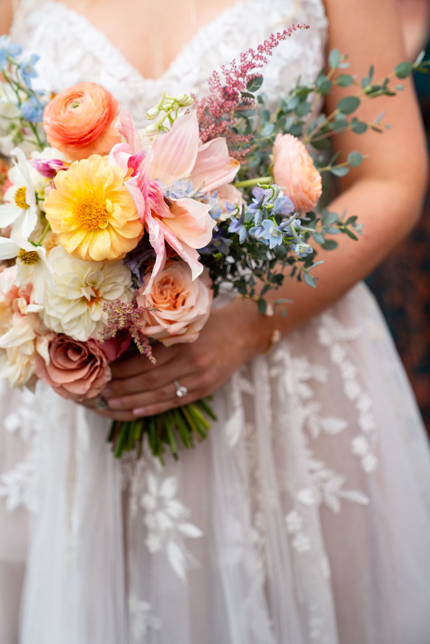 Close-up of bride holding a romantic spring bouquet with peach ranunculus, pink roses, yellow dahlias and eucalyptus greenery.
