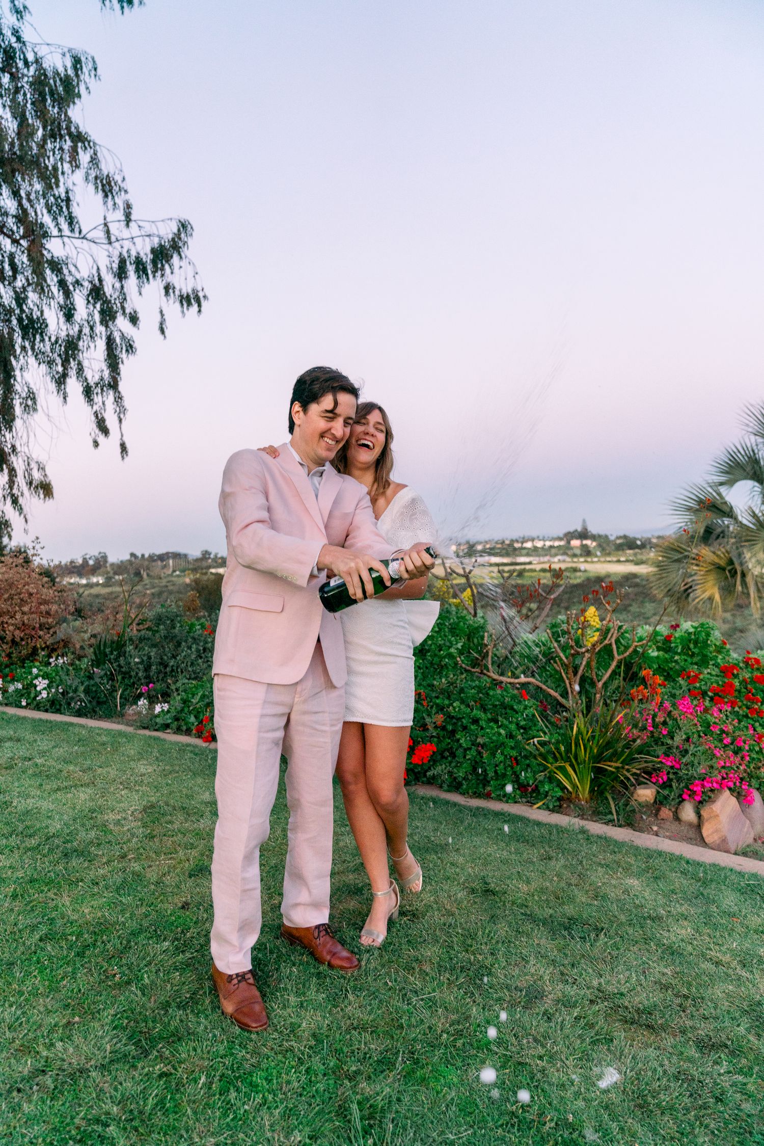 Couple celebrating with champagne in garden at sunset wearing pink suit and white dress standing on lawn.