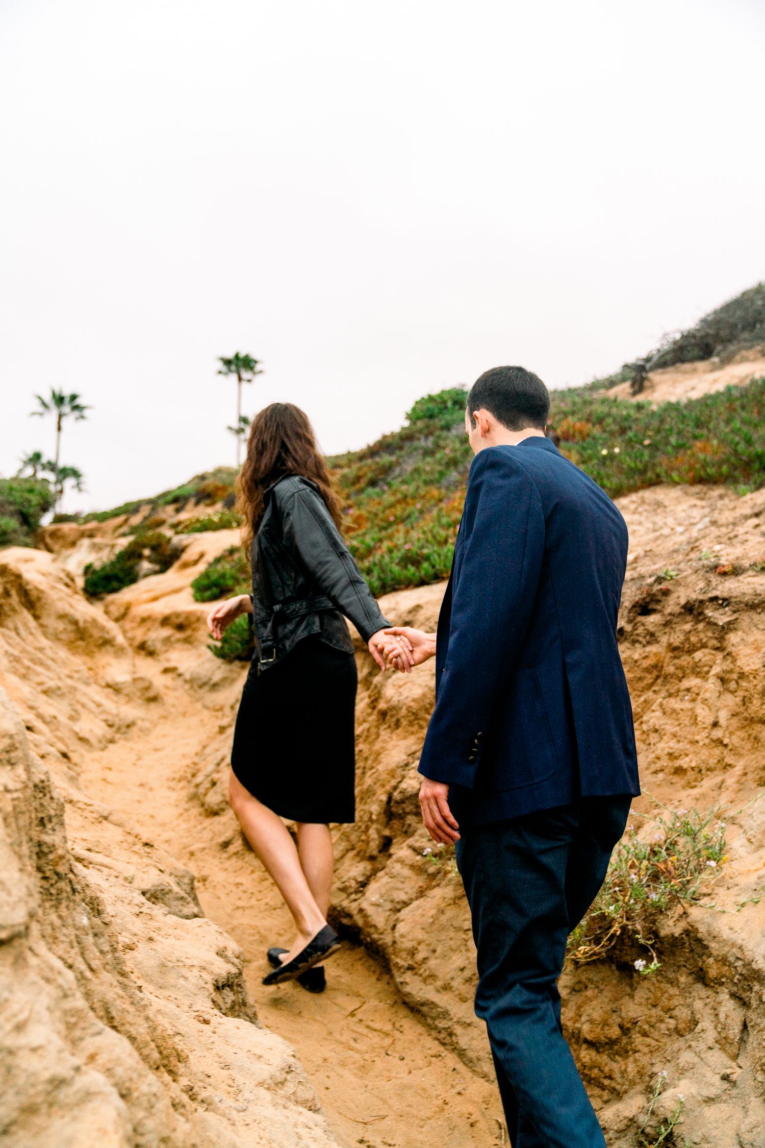 Two people walk together holding hands along a sandy coastal trail with palm trees and cliffs in the background.