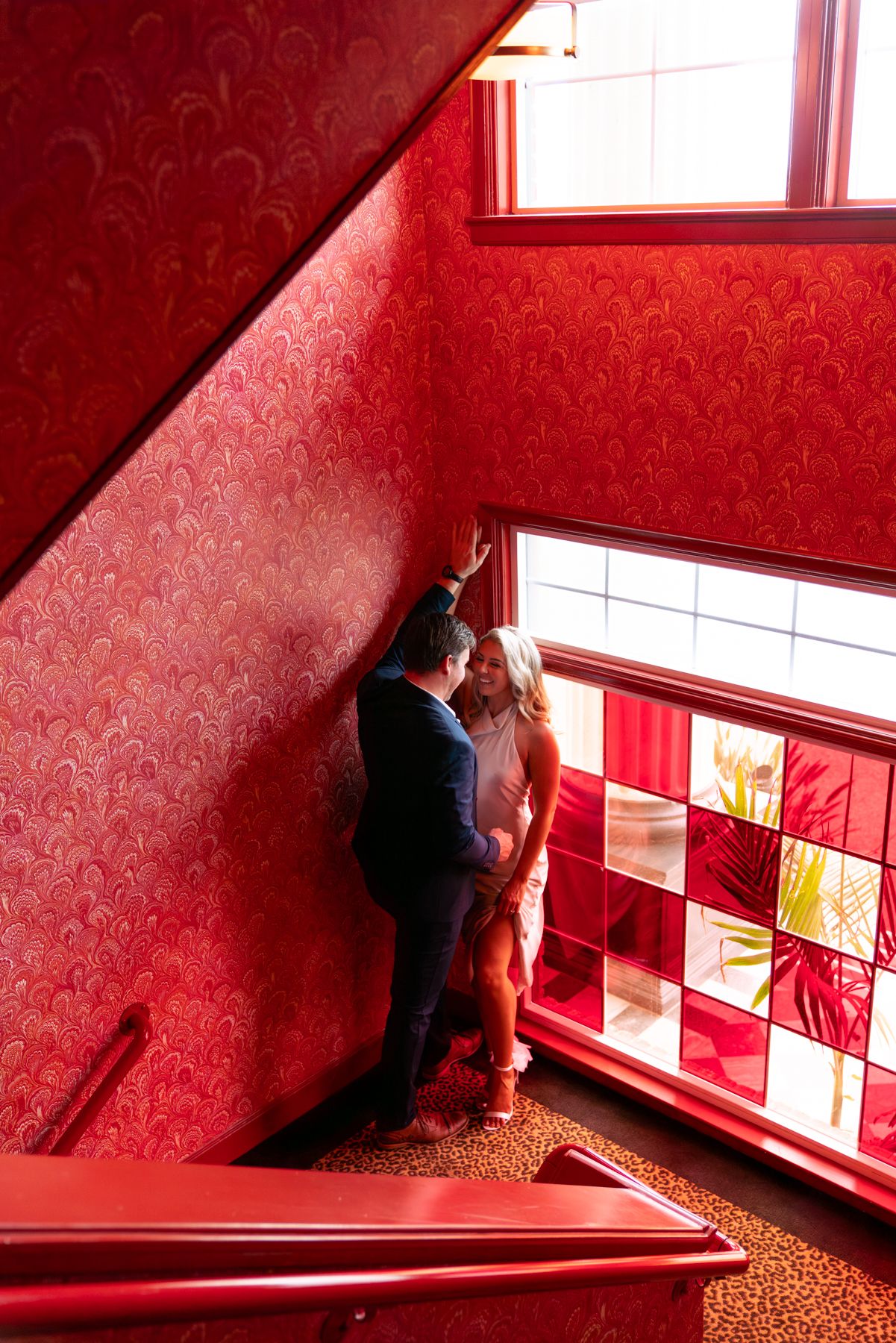 Two people stand in a stairwell with vibrant red damask wallpaper and a decorative stained glass window.