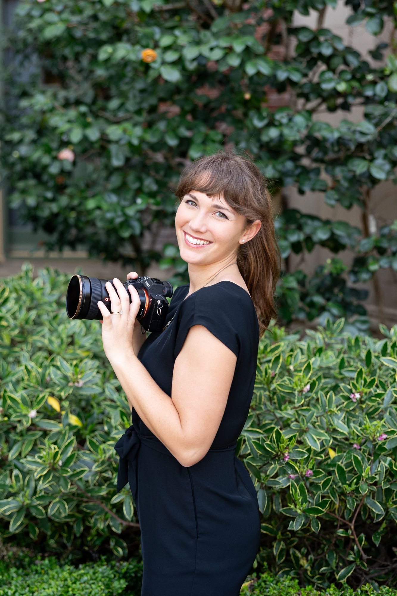 A photographer in a black dress holds a professional camera while standing in front of lush green foliage.