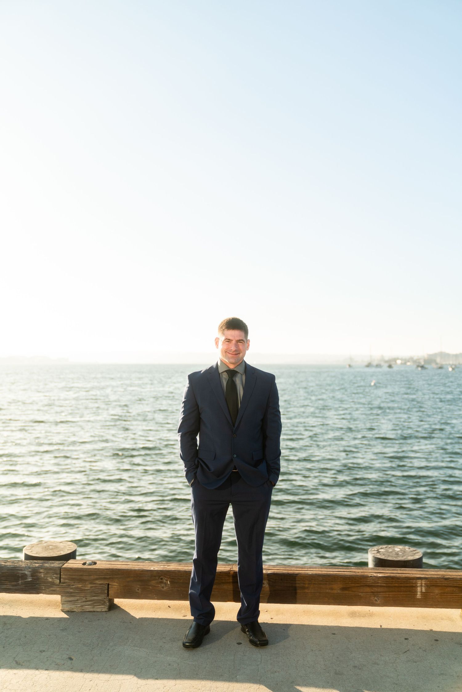 A cropped panoramic view shows someone standing at a concrete seawall overlooking calm ocean waters on a bright day.