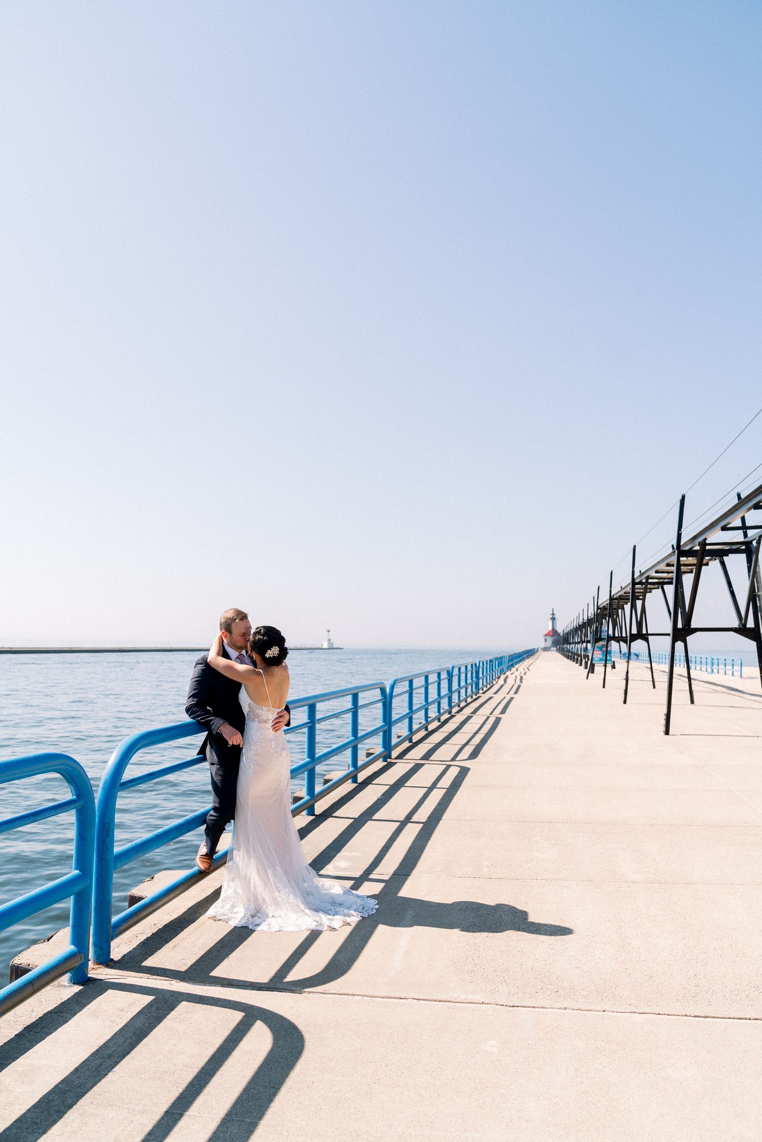 A couple embraces on a white concrete pier with blue railings along Lake Michigan on a sunny day.