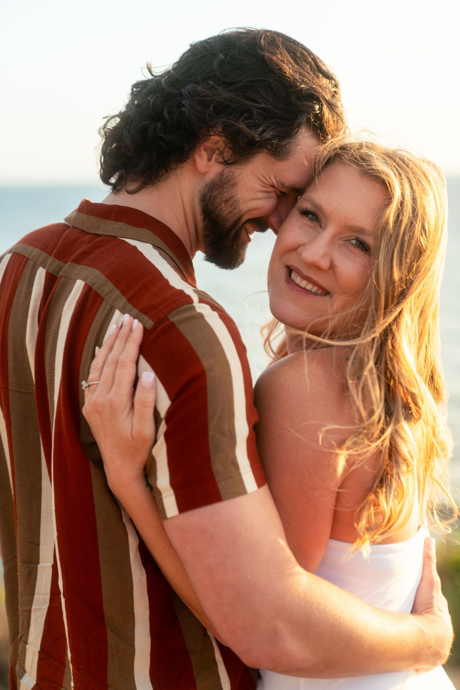 A smiling couple embraces during sunset, with golden light illuminating their intimate moment against a blurred background.