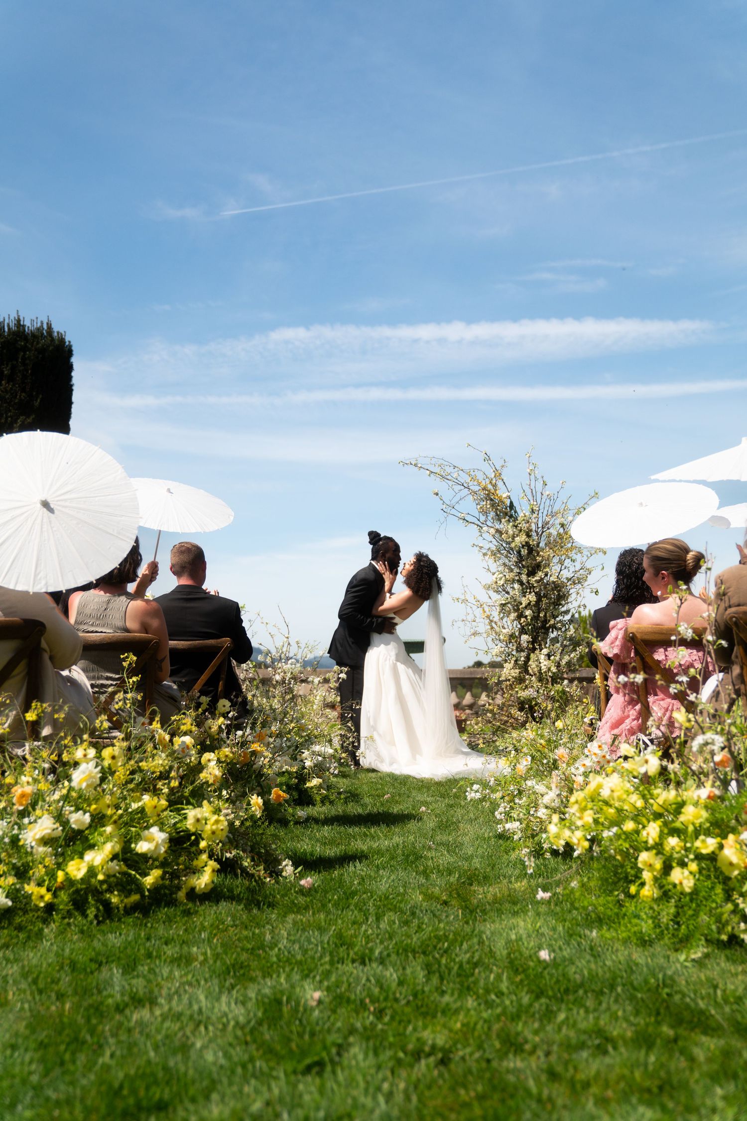 Wedding ceremony takes place outdoors on a grassy hill with yellow flowers and white parasols under blue summer sky.