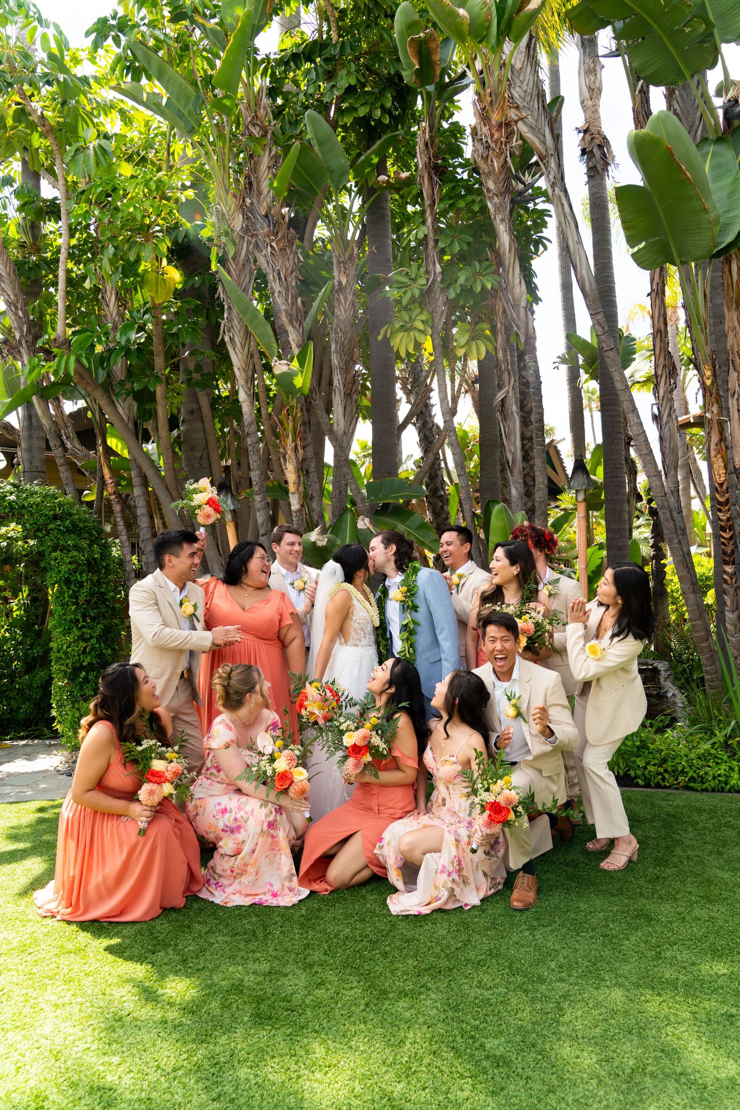 Wedding party in coral and neutral colored outfits posing on lawn with tropical palm trees in background.