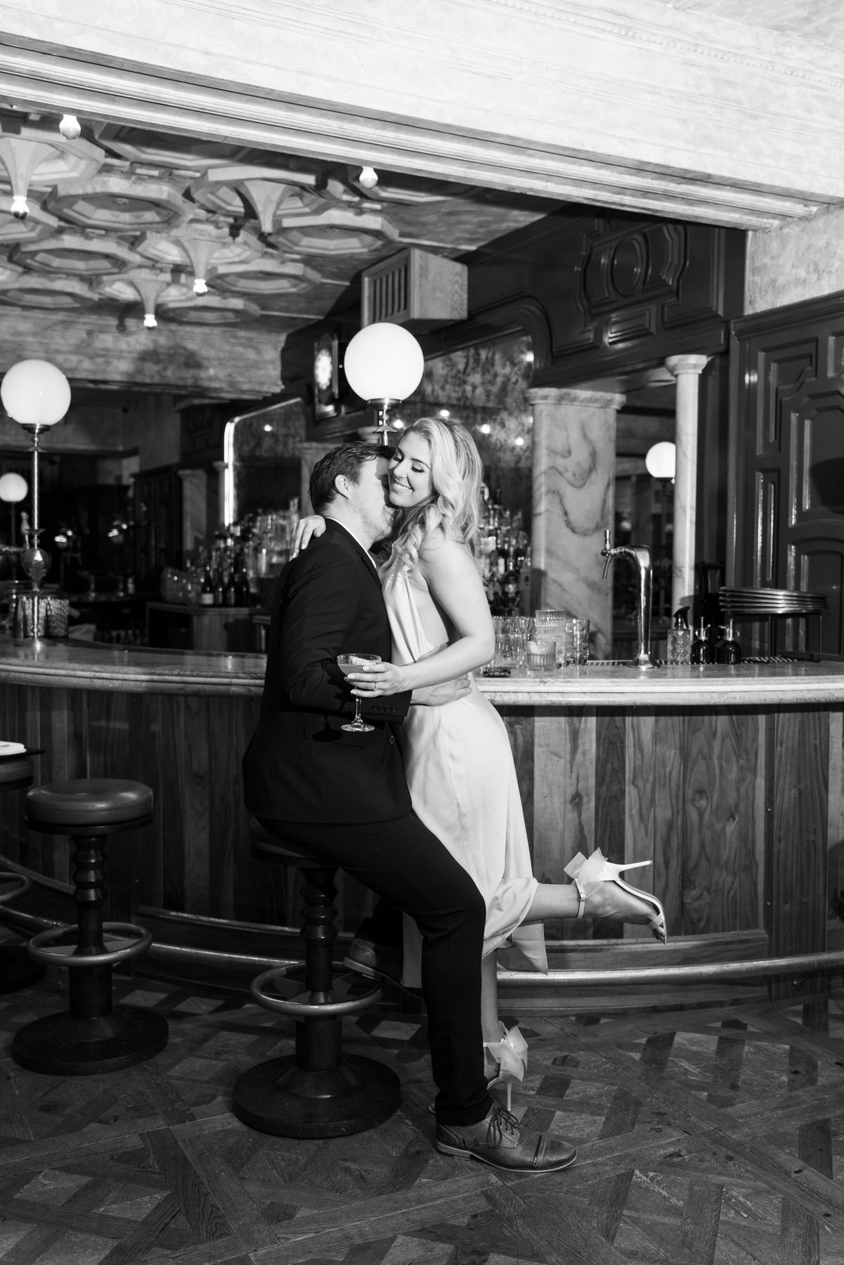 A black and white photograph of a couple in formal attire embracing at a vintage-style bar with round lights overhead.