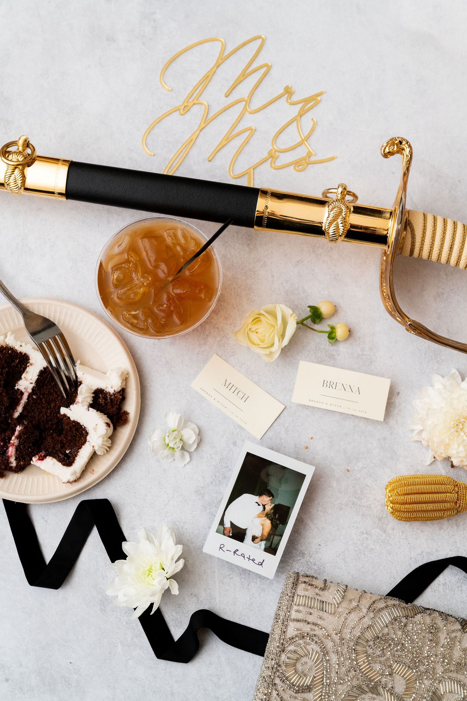 Wedding details flatlay with gold sword, black ribbon, chocolate cake, cocktail and polaroid photo on light gray surface.