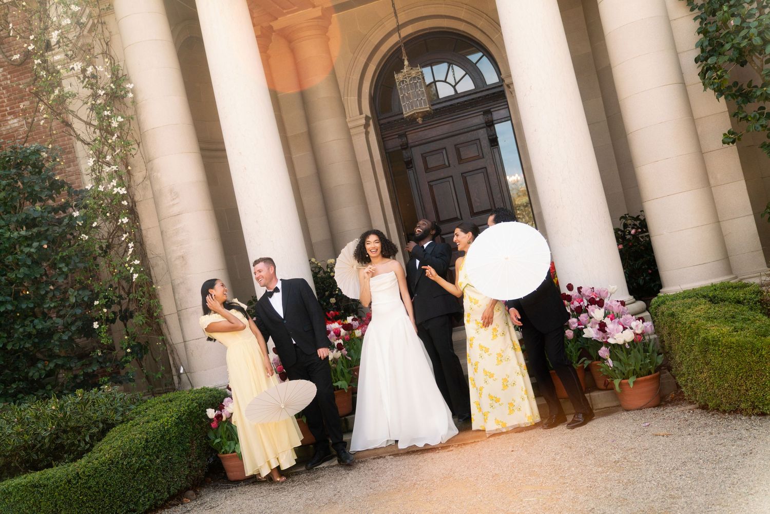 A wedding party gathers outside an elegant columned entrance with floral decorations and manicured hedges.