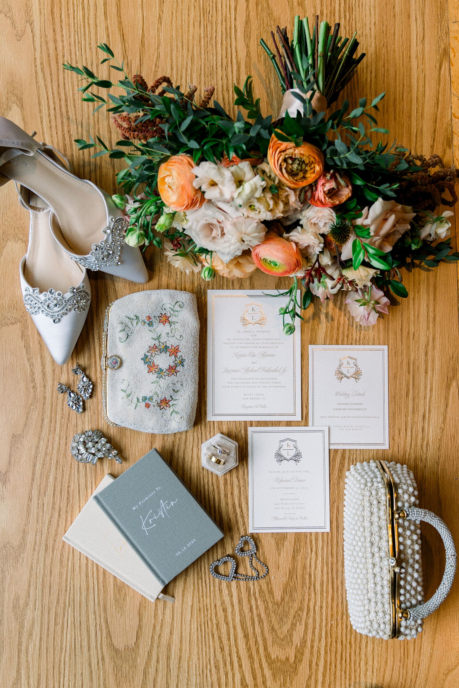 Wedding accessories flat lay with floral bouquet, invitation suite, pearl clutch and embellished shoes on wood background.